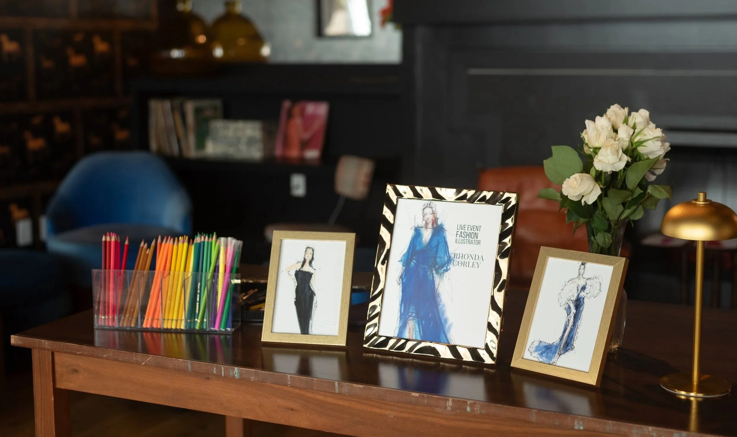 A wooden table with a container of colored pencils, three fashion illustrations in framed art, a vase of white roses, and a small gold desk lamp. In the background, there is a dark wall with books and a blue chair.