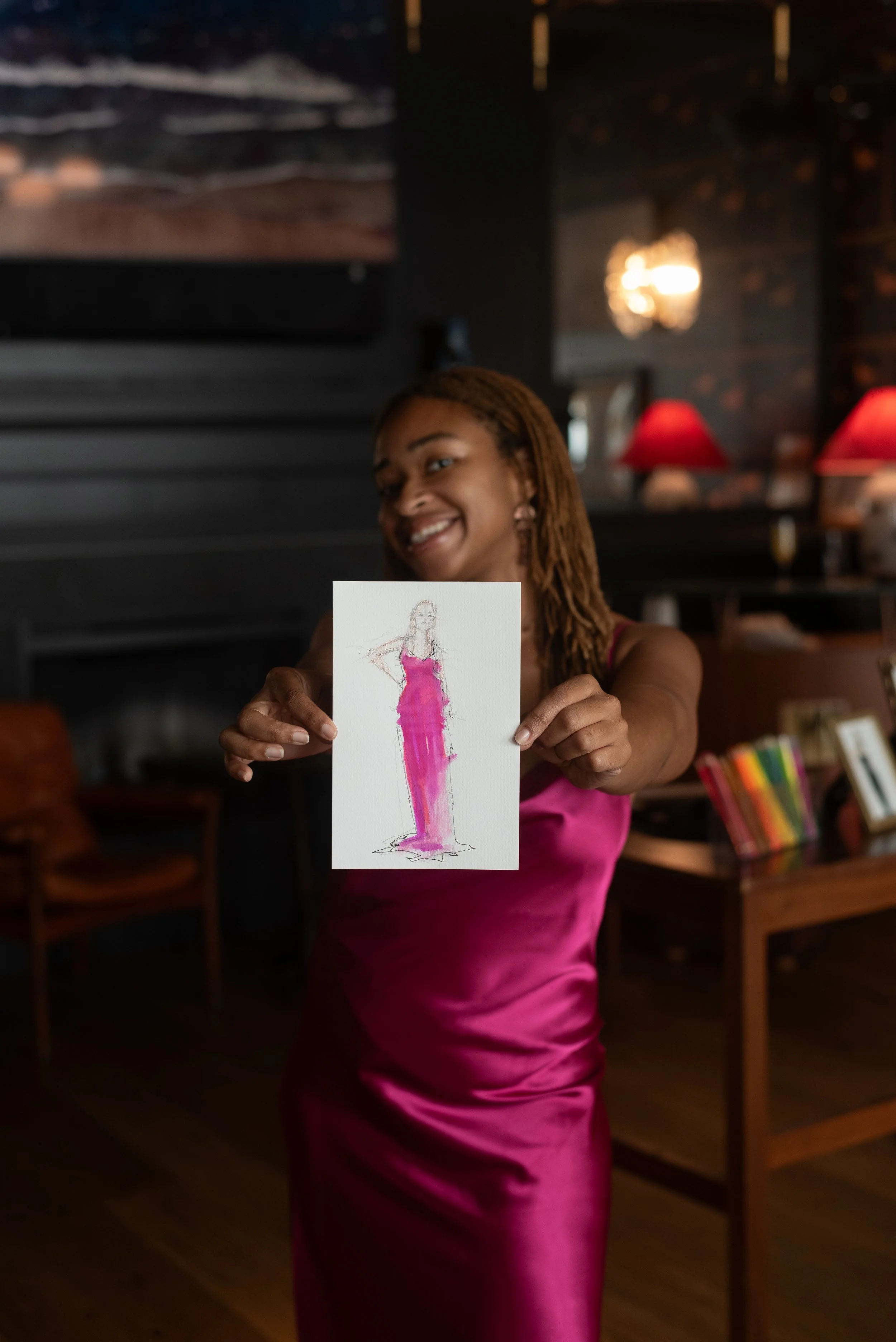A woman in a pink satin dress holding a small drawing of herself wearing a pink dress, smiling in a dimly lit interior with red lamps and dark wood decor.
