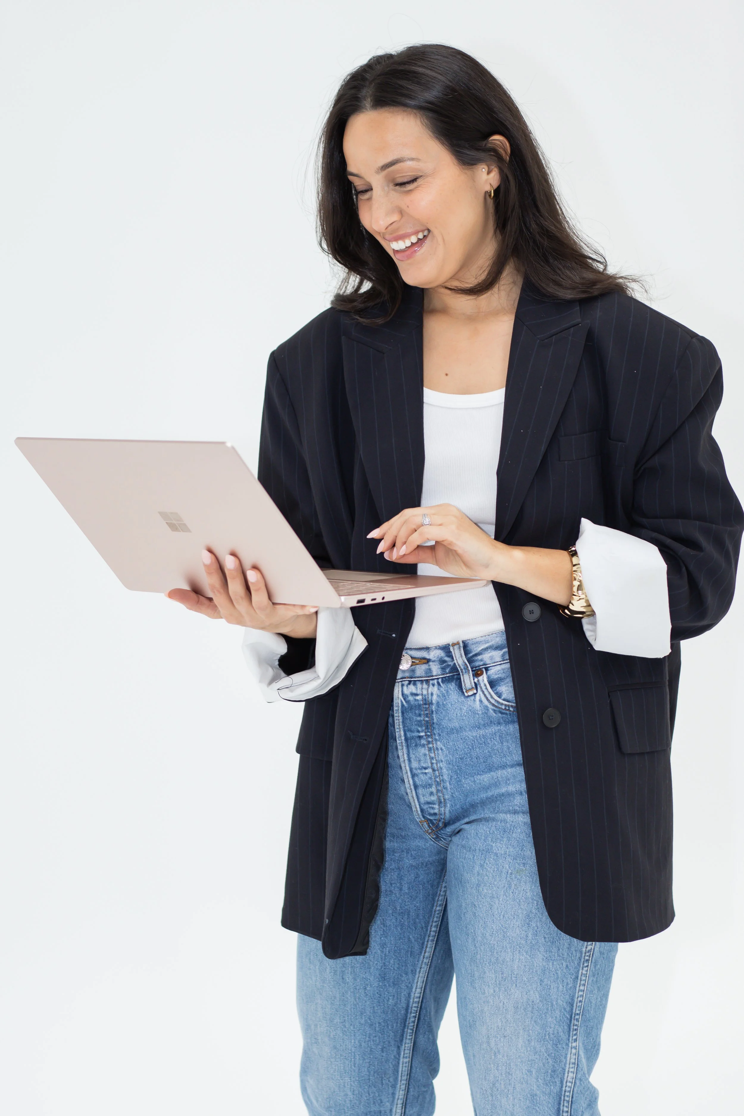 A smiling woman with dark brown hair, wearing a black pinstripe blazer over a white shirt, and blue jeans, is looking at a rose gold Microsoft Surface tablet.