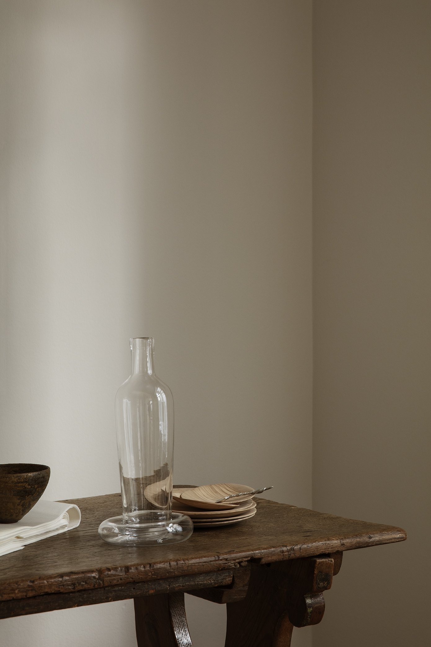 Empty wooden table with a glass bottle, a dark bowl, a stack of small plates, and a fork.