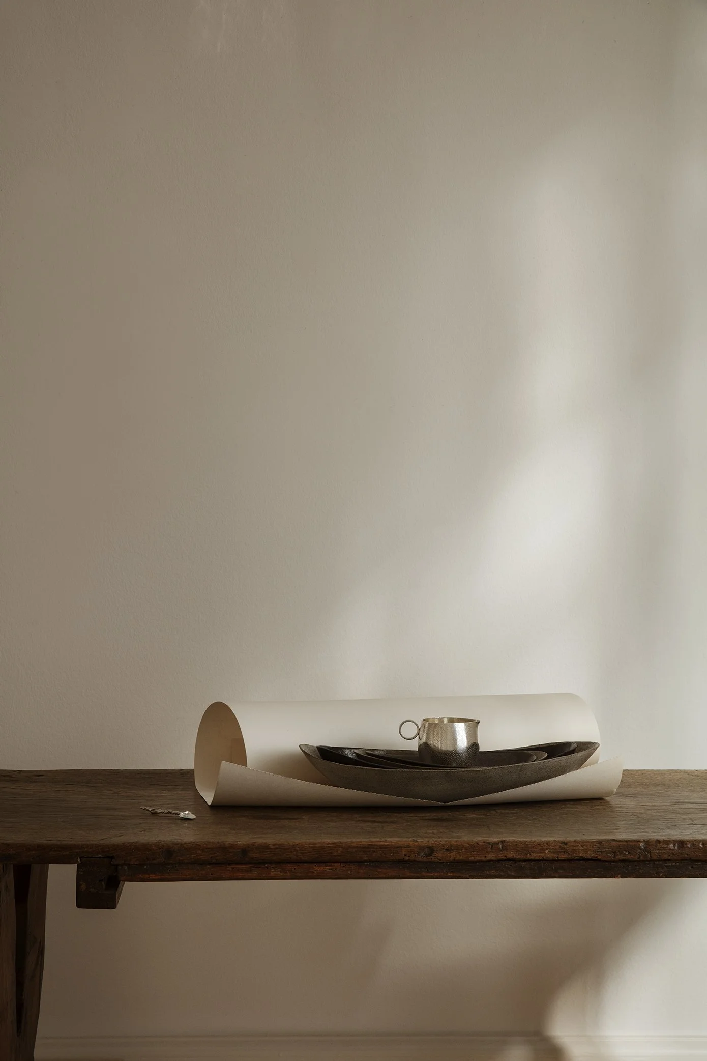 A wooden table with a rolled-up sheet of paper, a black plate, a small metallic cup, and a black tray arranged on it, against a plain light-colored wall.