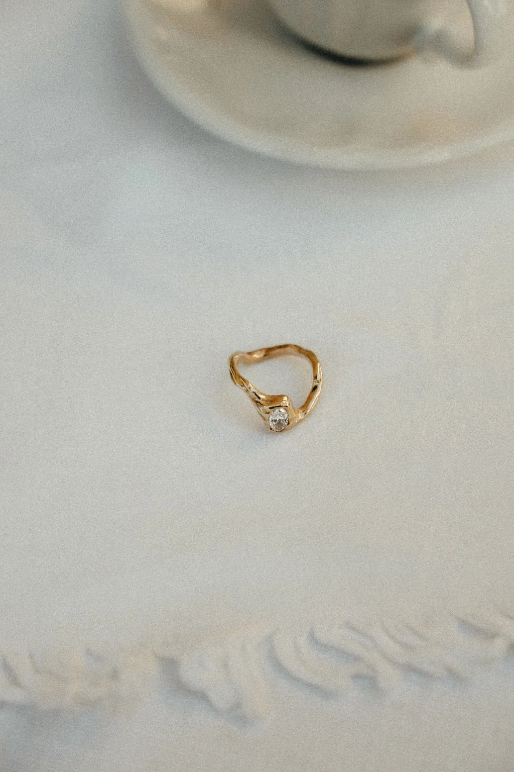 Close-up of a gold ring with a small diamond, placed on a white surface near the edge of a white cup placed on a saucer.