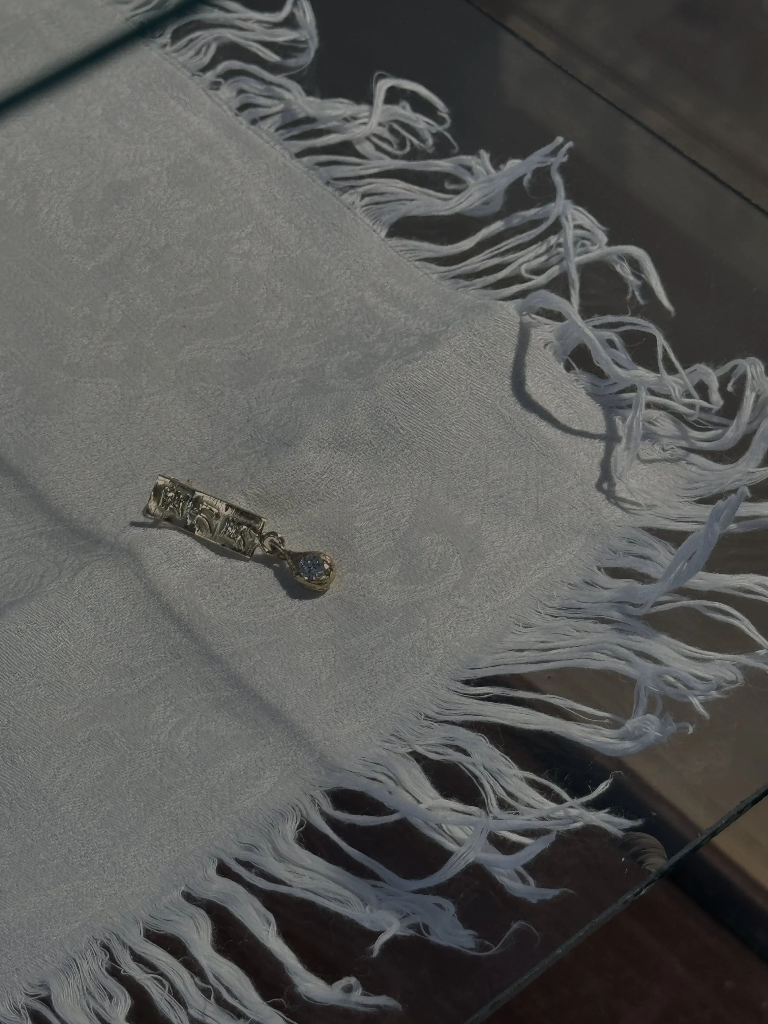 A silver pendant with a small blue gemstone on a white textured cloth with fringes, placed on a wooden surface.