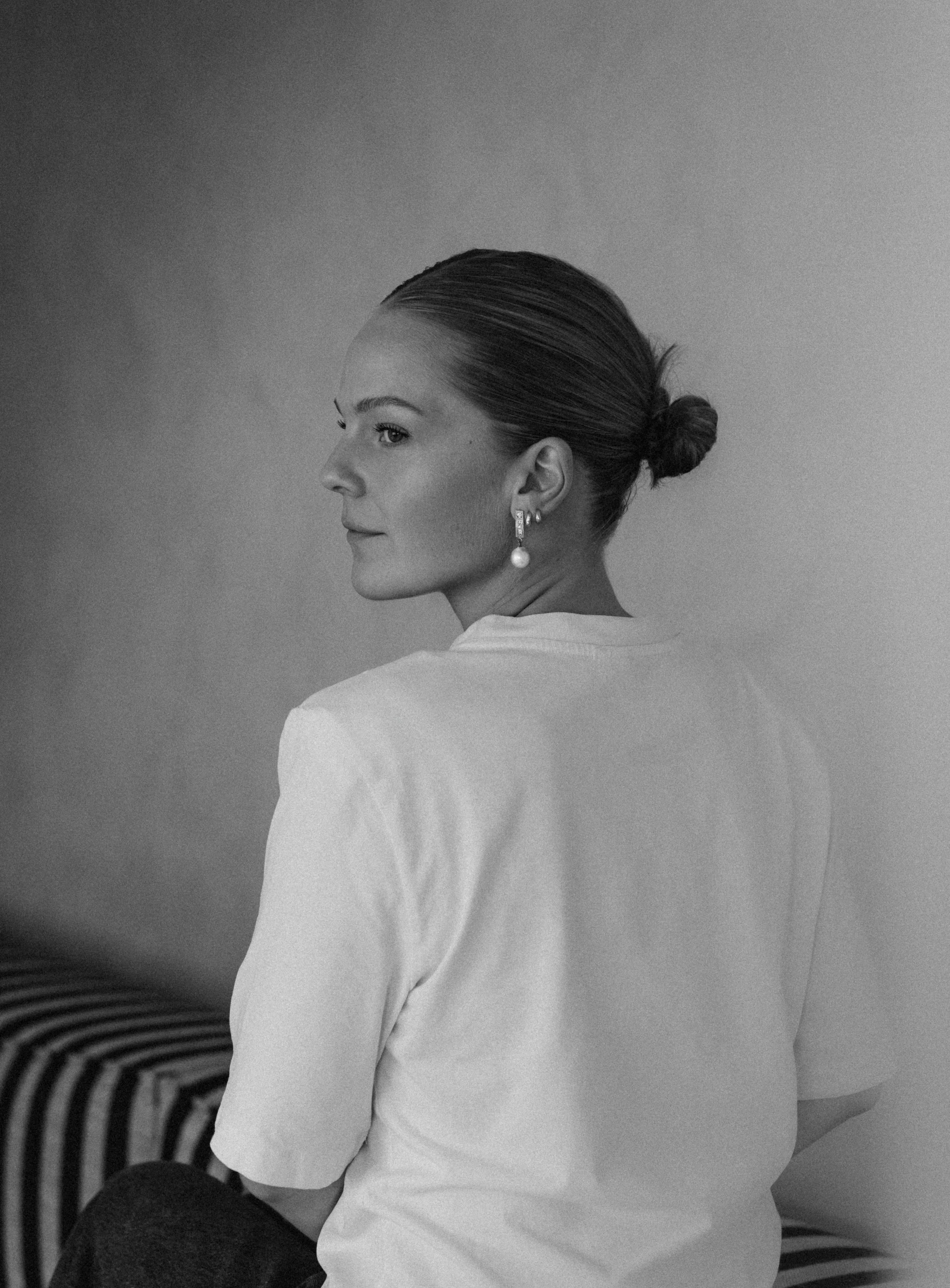 Black-and-white photo of a woman with her hair in a bun, wearing earrings and a light-colored top, looking to the side, sitting against a plain background.
