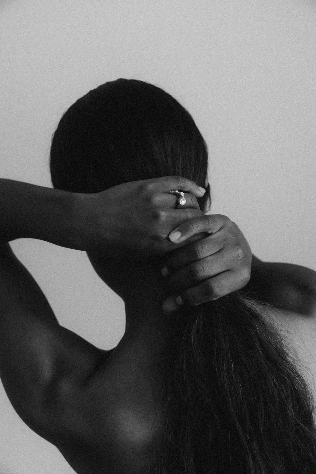 A black-and-white photo of a person with long hair covering their face, with one hand resting on their head and the other on their neck, wearing a ring on their finger.