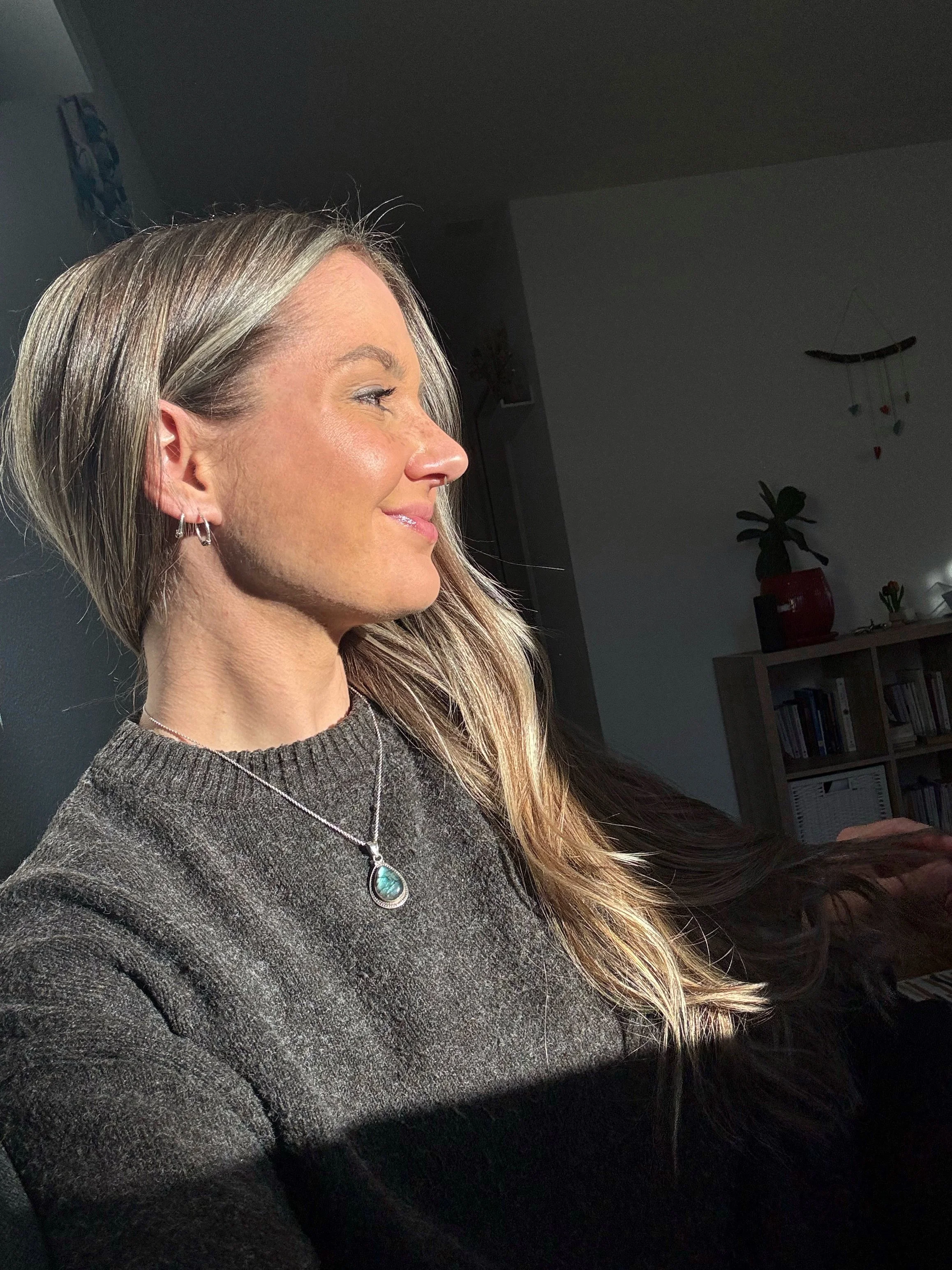 Femme avec des cheveux gris et longs, portant un collier argenté avec une pierre verte, souriant en regardant vers la gauche, éclairée par la lumière naturelle.