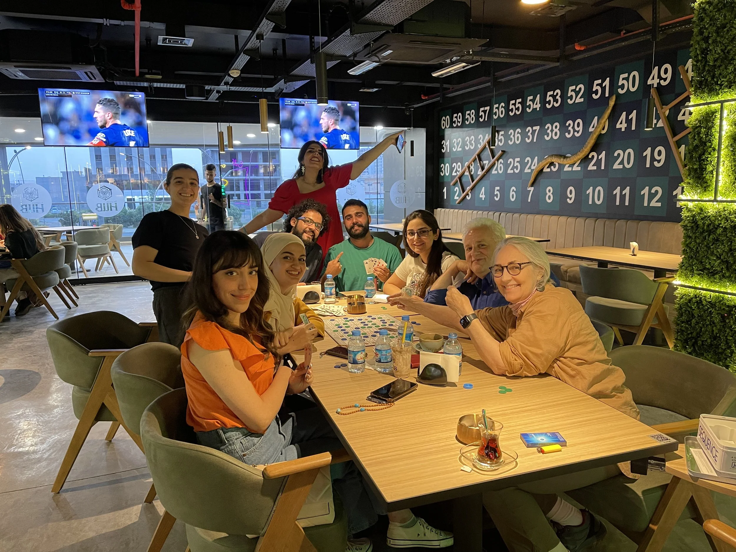 Group of nine people sitting around a table inside a restaurant, with two women standing, one in a red top, the other in a black top. All are smiling, with drinks and playing cards on the table. Two television screens display a soccer player in the background, and a large wall with seating chart numbers is visible in the background.