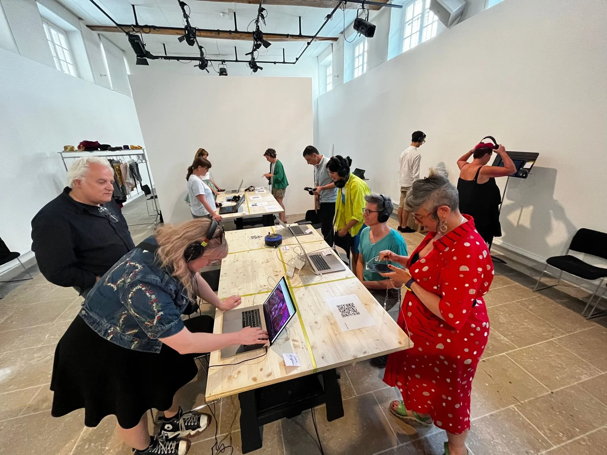 People gathered around a long wooden table with laptops, headsets, and smartphones in a bright, high-ceiling room with large windows. Some individuals are standing, some are sitting, and one woman is wearing a red polka-dotted dress.