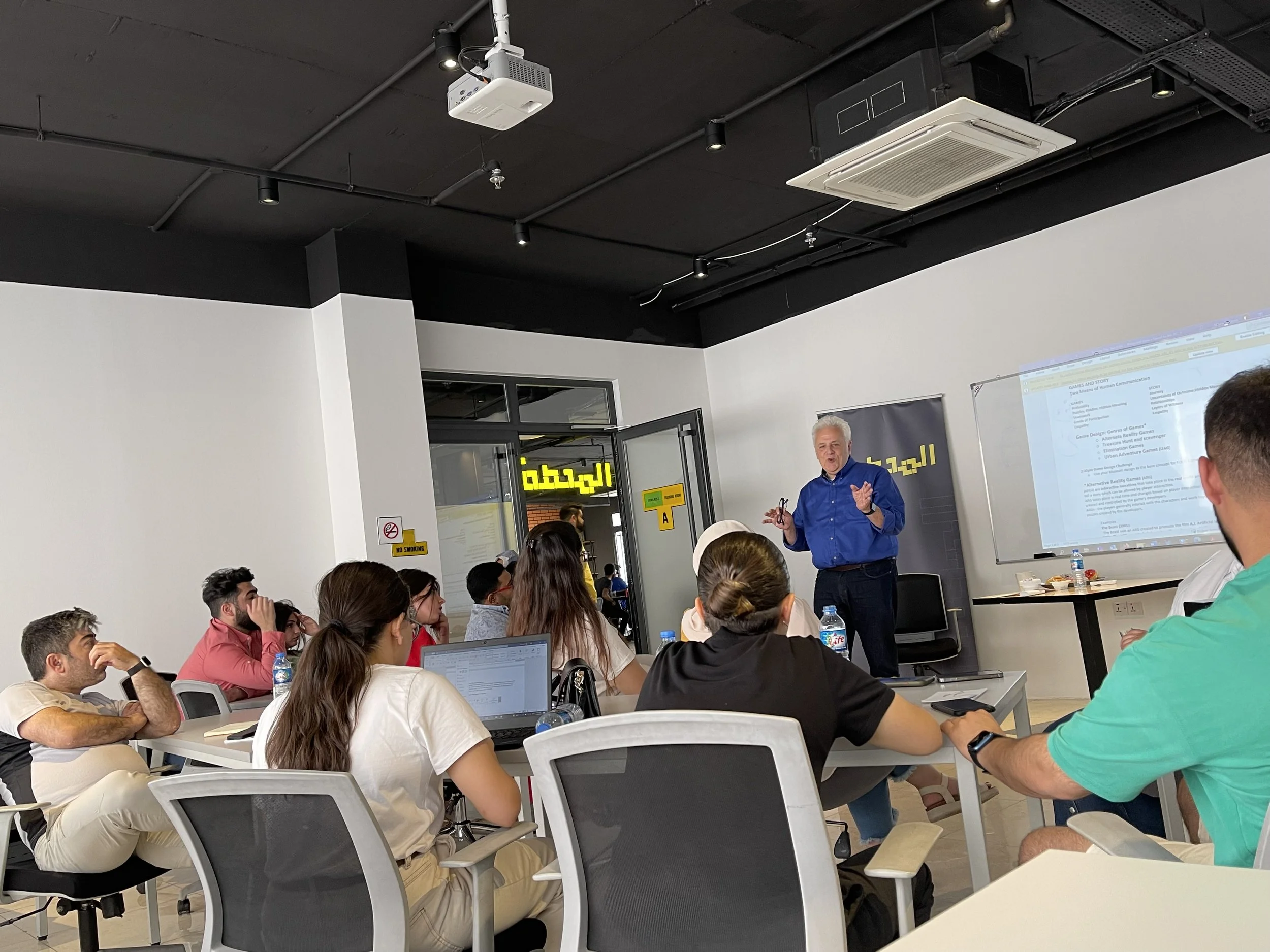 A man in a blue shirt standing in front of a classroom giving a presentation to students seated at tables, some with laptops, in a room with a high ceiling, black ceiling, and walls, with a white projection screen and various signs.
