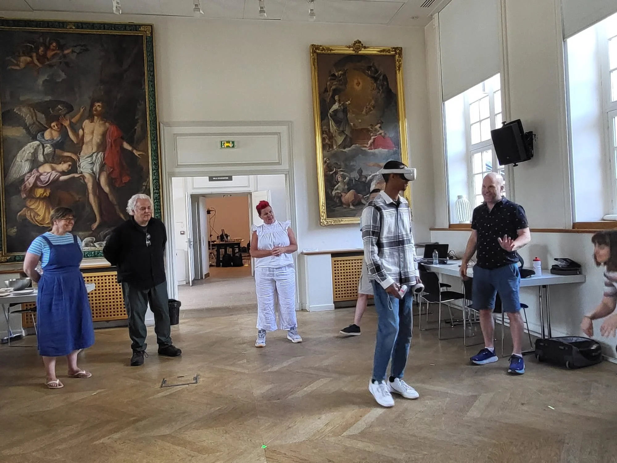 People in a gallery or museum, with classical paintings on the wall, some standing and some engaging with virtual reality headsets. The room has high ceilings, large windows, and wooden flooring.