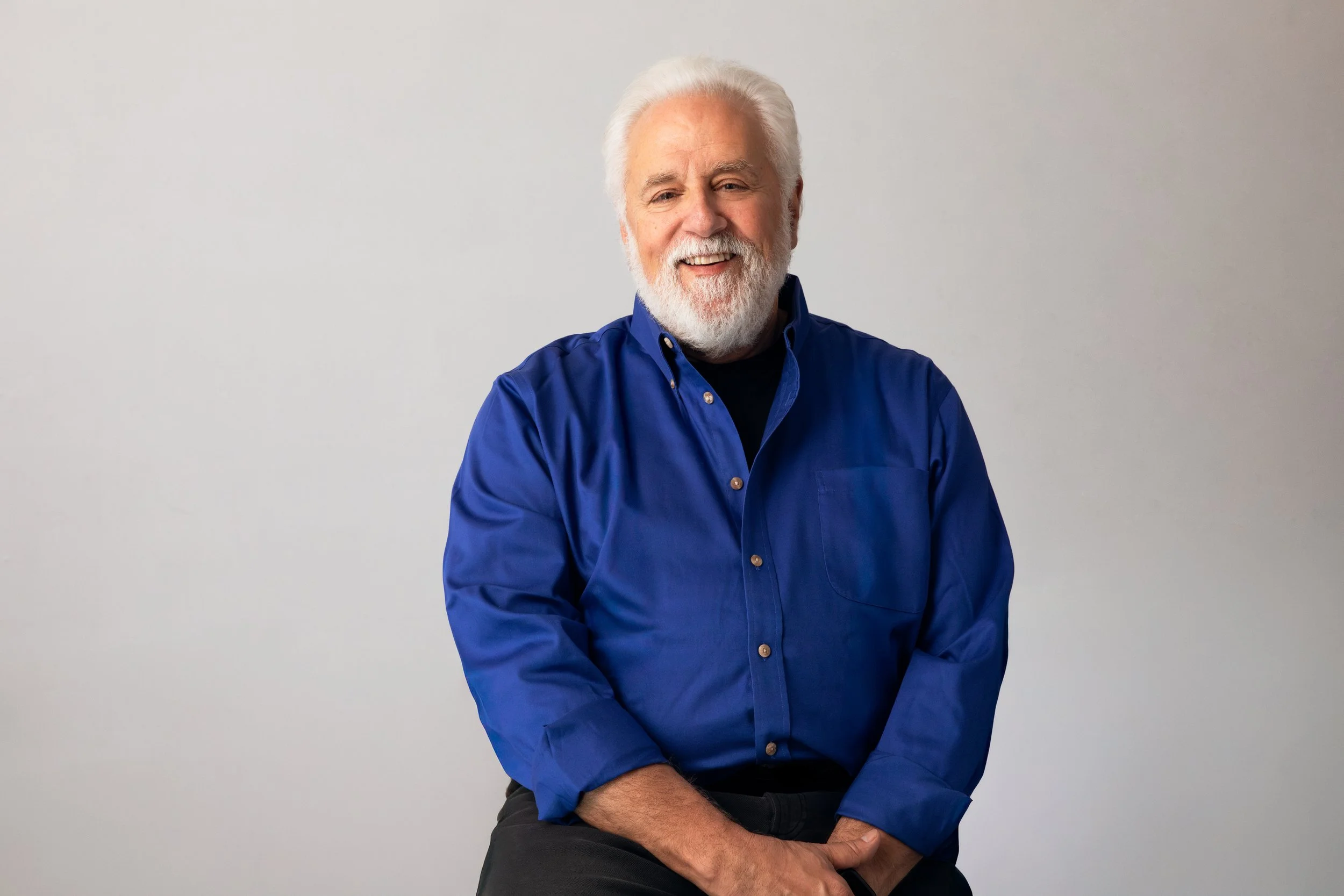 An elderly man with white hair and a beard, smiling, wearing a blue button-up shirt, sitting against a plain light background.