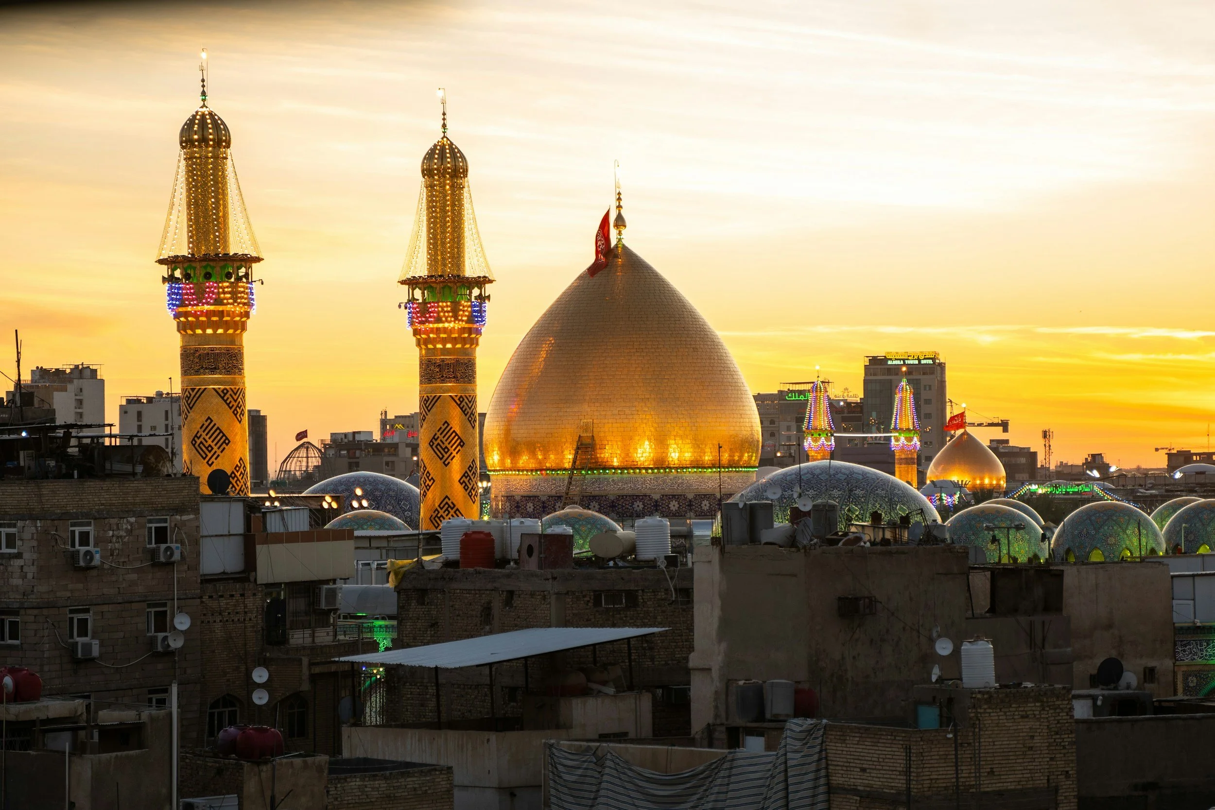 Cityscape at sunset featuring a mosque with golden domes and two tall minarets decorated with lights, surrounded by various buildings.