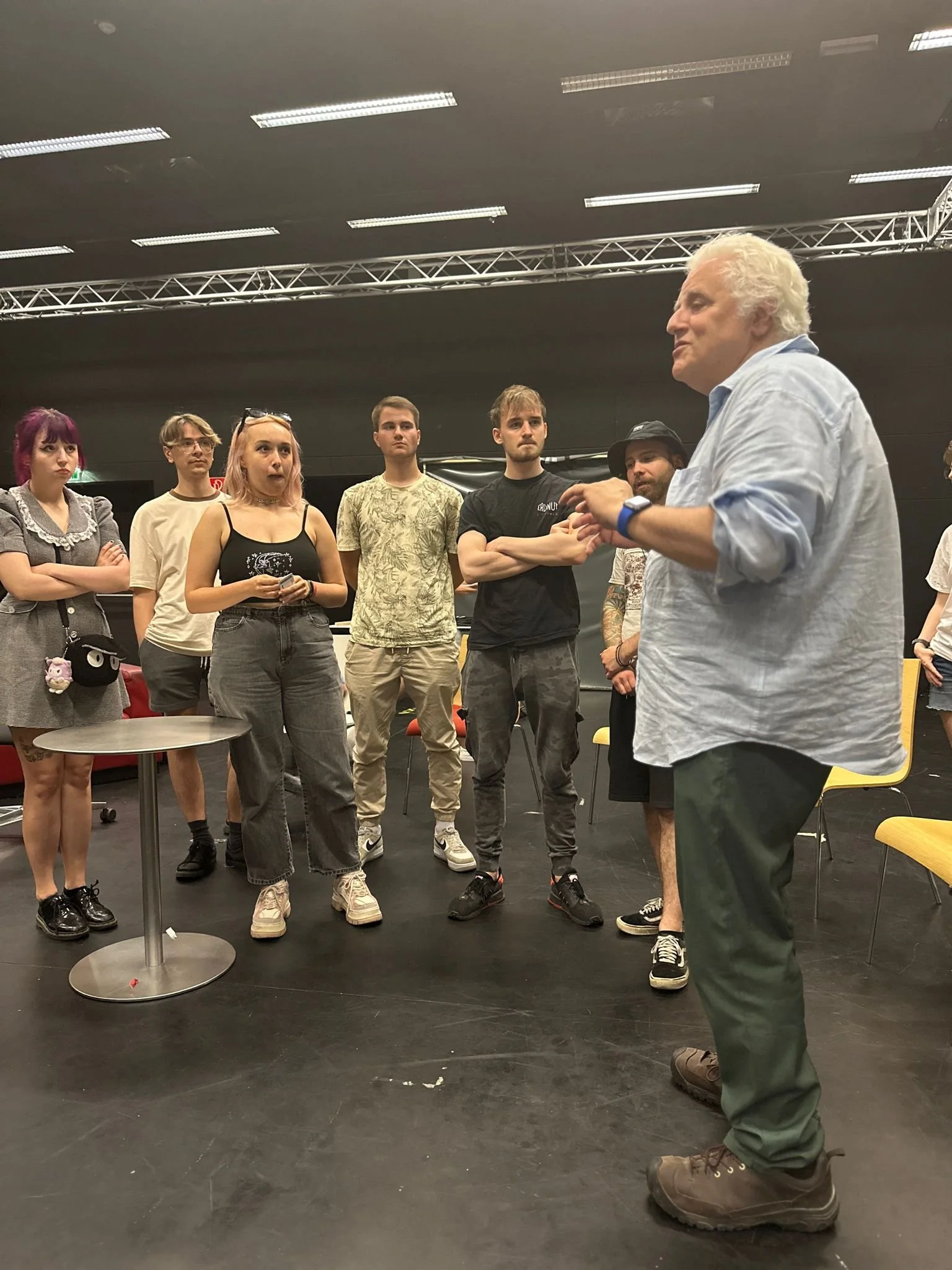 An older man with white hair and a light blue shirt speaking to a group of young adults in a room with black walls and ceiling lights.