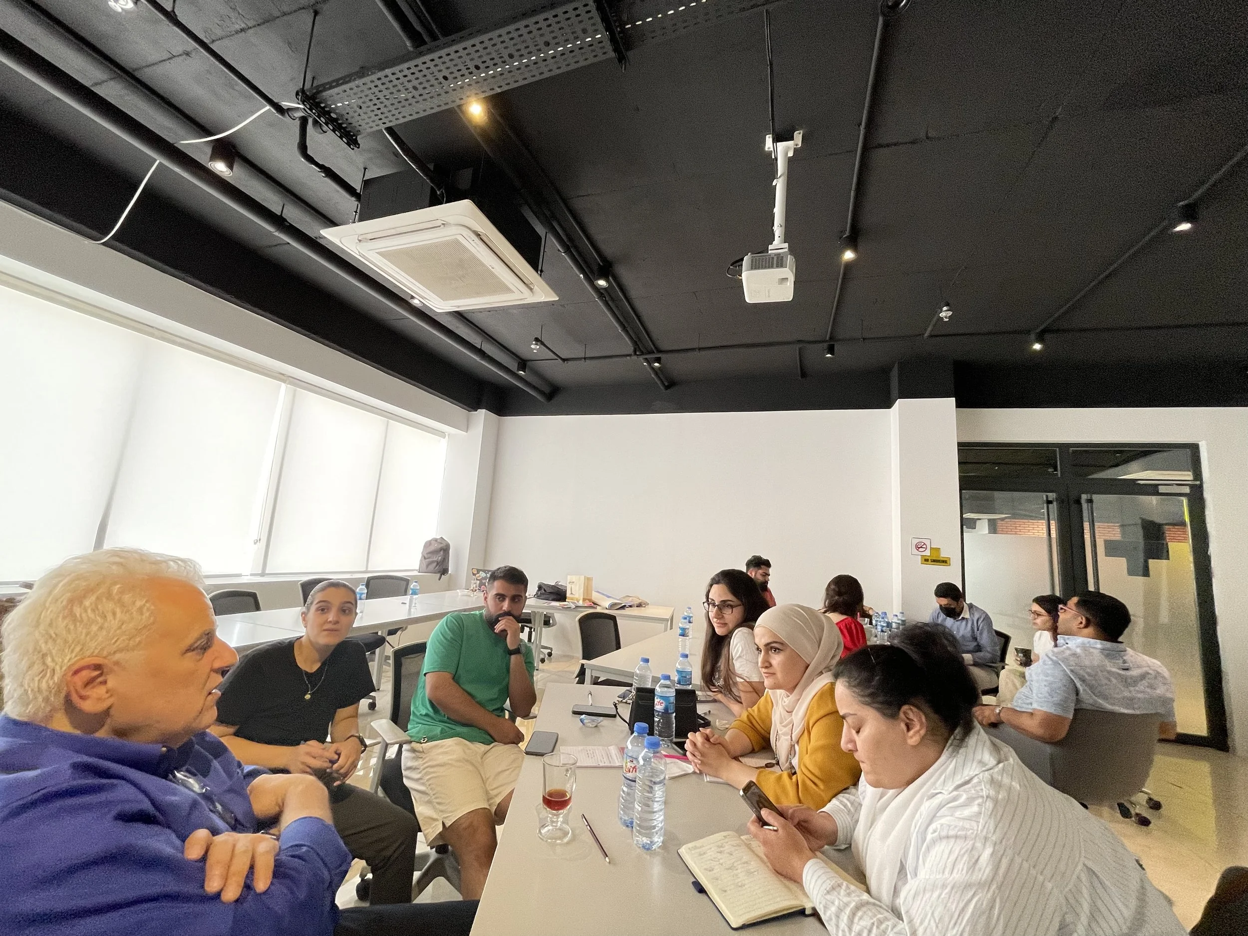 A diverse group of people sitting around a large conference table in a modern meeting room, engaging in a discussion or meeting. There are notebooks, water bottles, and glasses on the table, and a ceiling-mounted projector and air conditioning unit are visible.