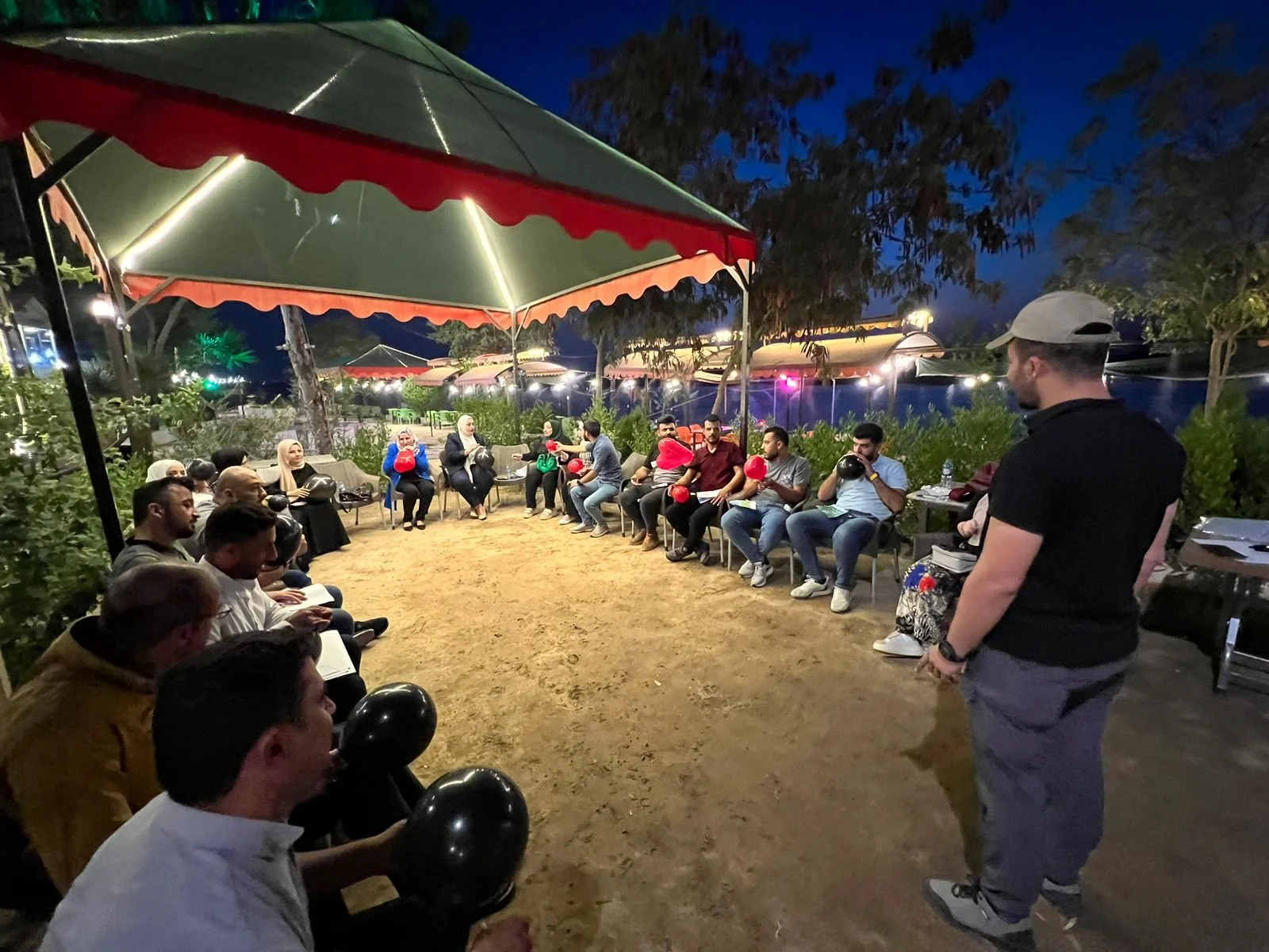 People sitting outdoors at night under umbrellas, participating in an event with balloons, while a man stands in front of them.