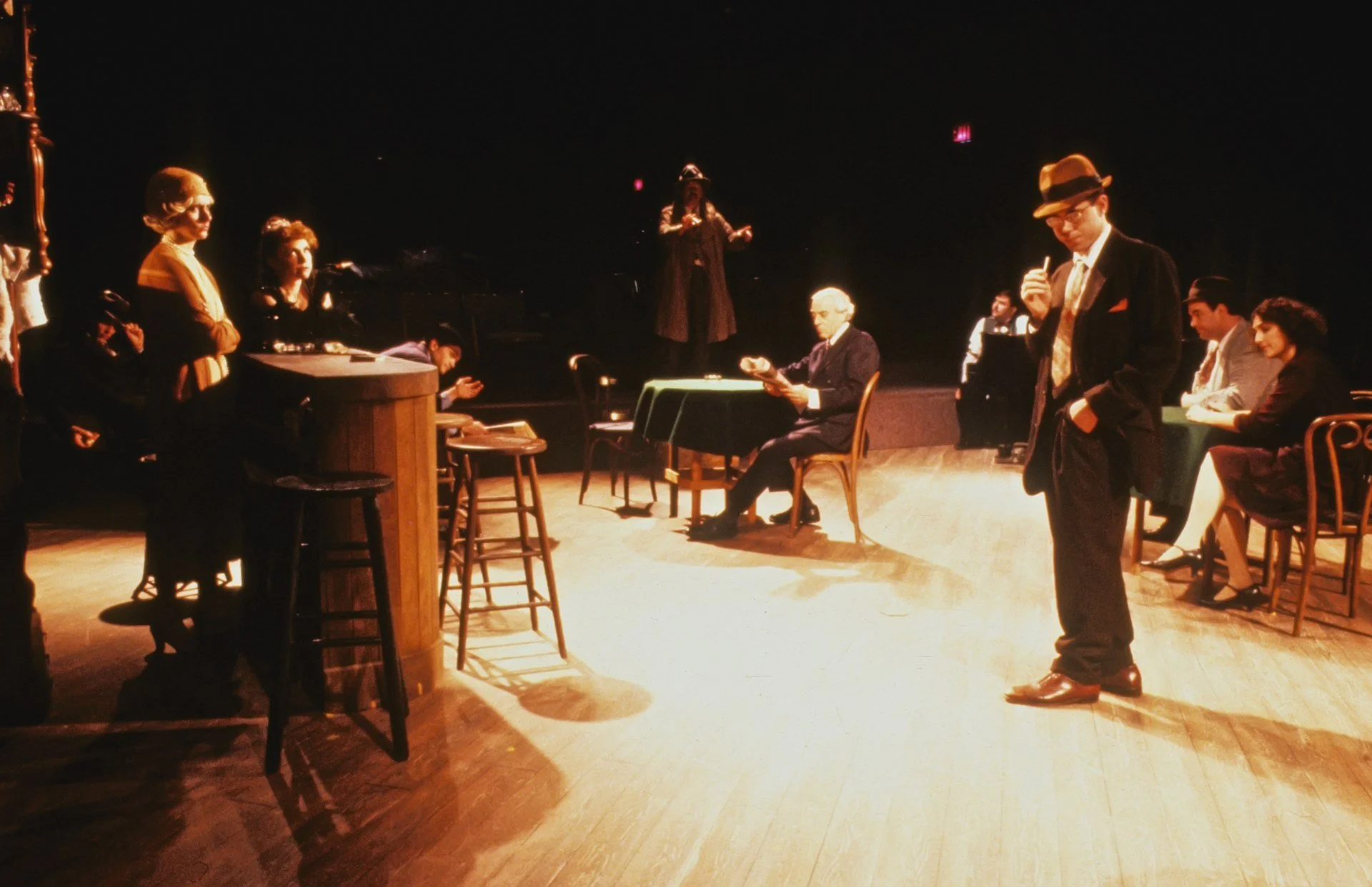 Scene from a theatrical play with multiple actors on stage, including a man in a suit and fedora reading a paper, a woman standing with crossed arms, a man sitting at a table, and a woman sitting and smiling. A performer is in the center, speaking or singing.