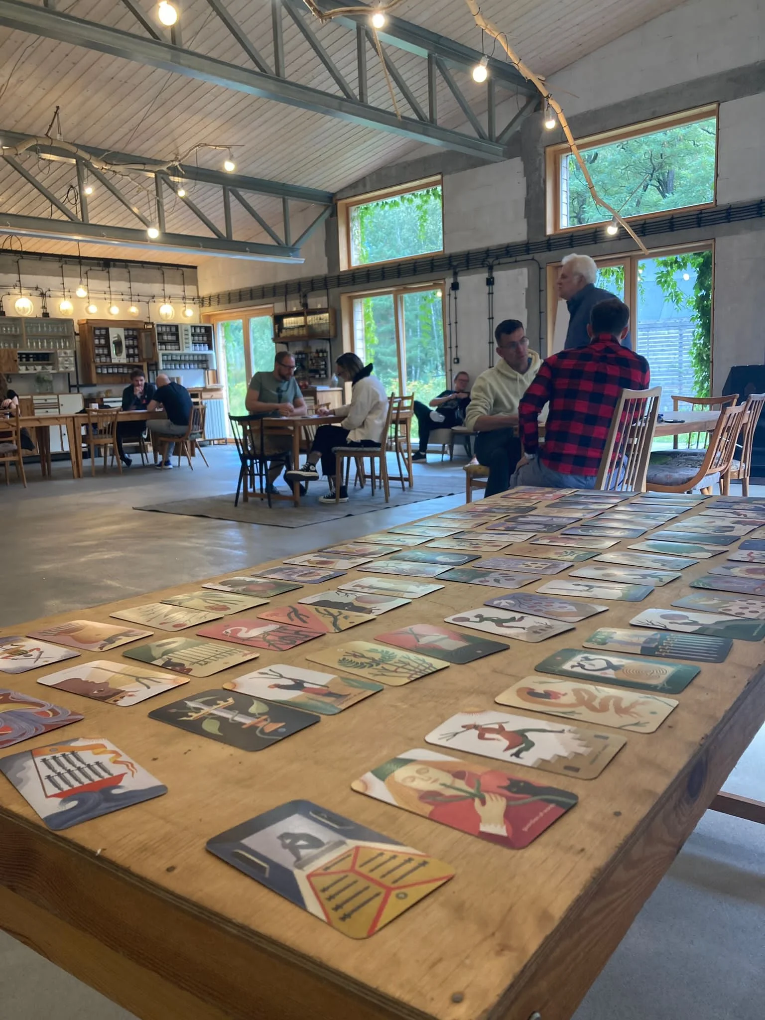 A group of people sitting at tables in a spacious room with large windows, wooden furniture, and string lights, playing a card game with illustrated cards laid out on a wooden table in the foreground.