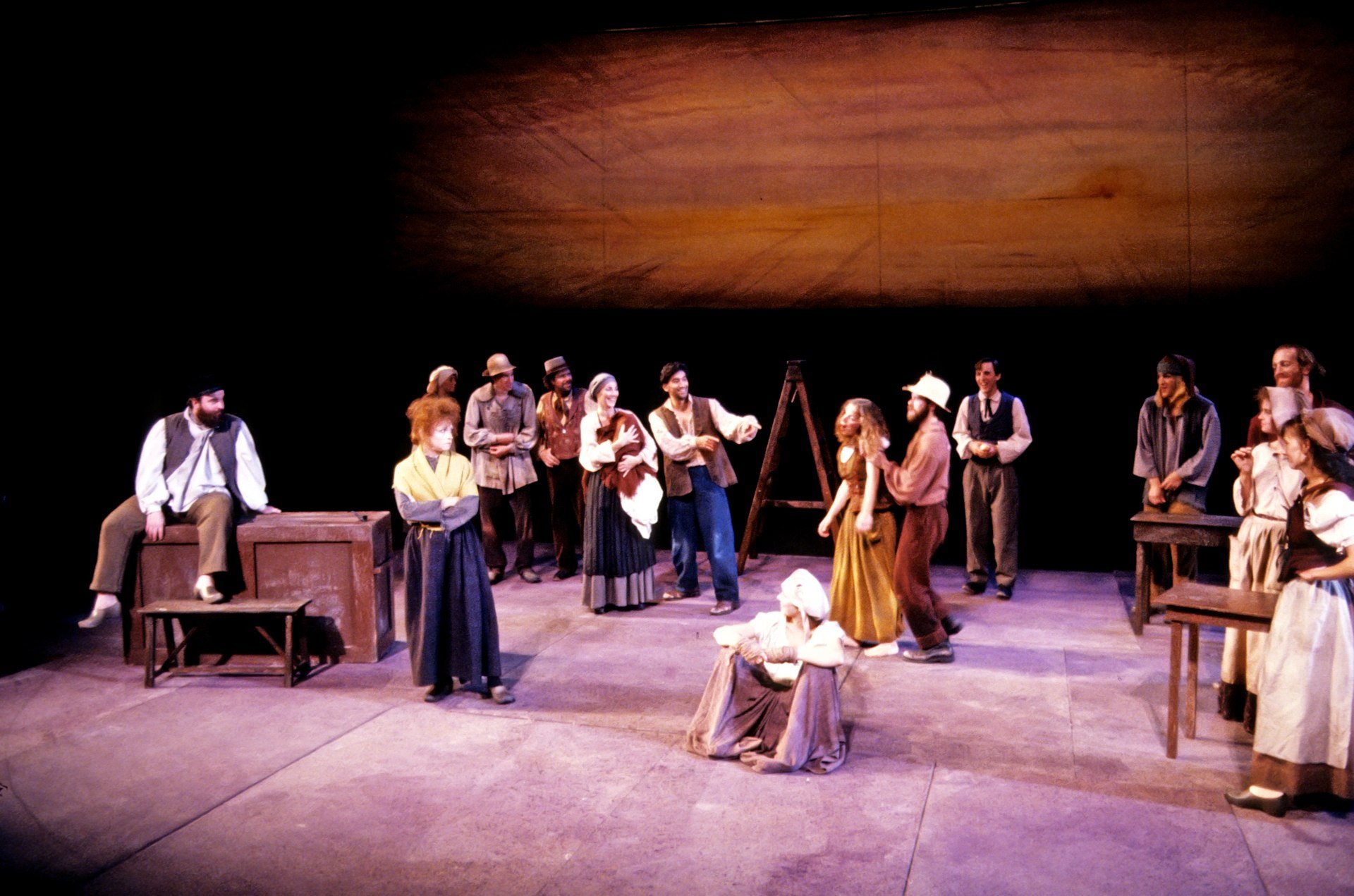 The image shows a group of actors on stage performing a play. They are dressed in period costumes, and some are standing while others are seated. The background features a large, plain wooden ceiling or backdrop, and stage props such as benches and a ladder are visible.