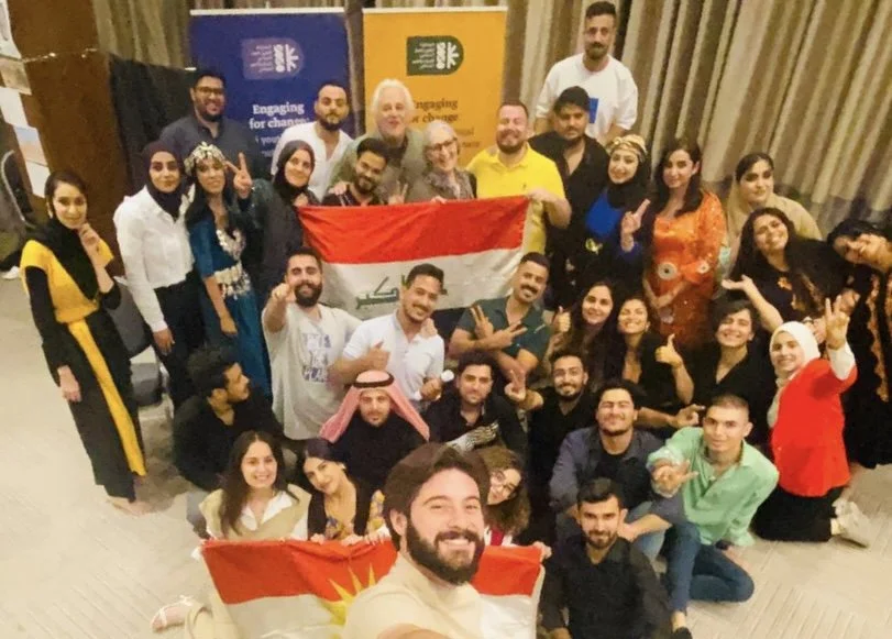 Group of people celebrating, some holding Kurdish and Iraqi flags, indoors with banners in the background.