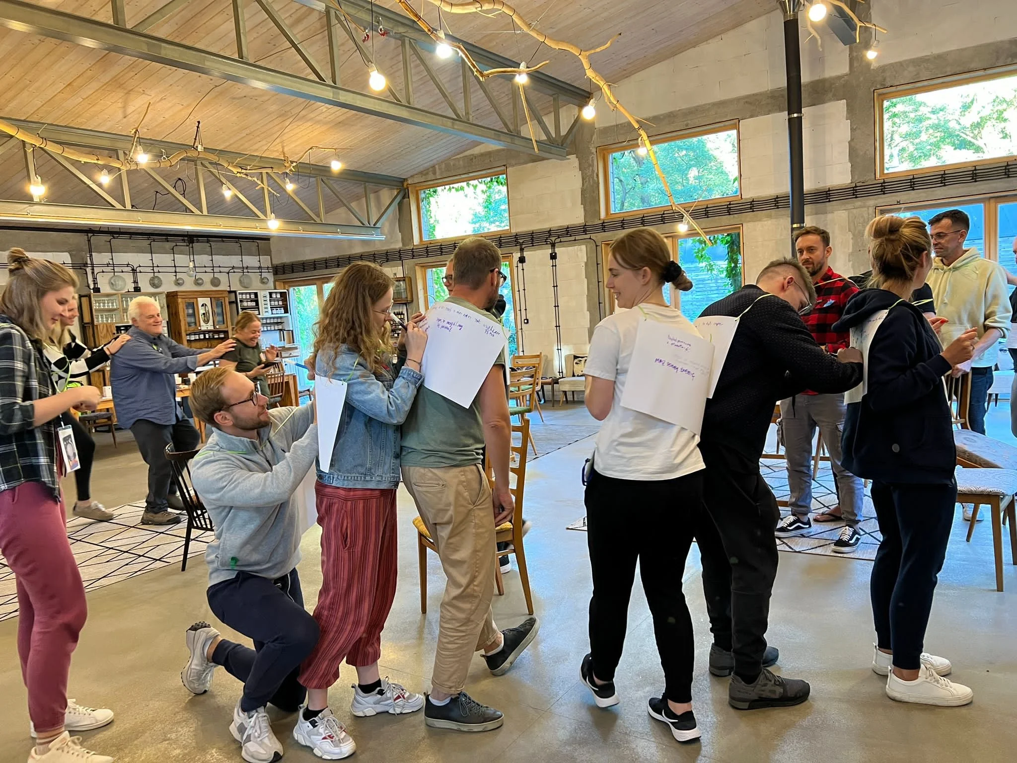 A group of people indoors, some standing and some kneeling, some holding papers or notebooks, engaging in a discussion or activity in a spacious, well-lit room with large windows, industrial decor, and string lights hanging from the ceiling.