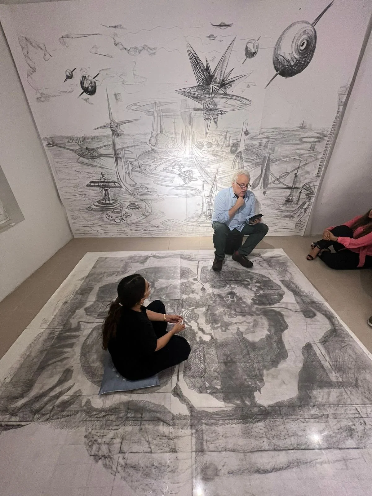 An art gallery with a large futuristic drawing on the wall and a floor-sized sketch on the floor. A man is sitting on a small stool reading from a tablet, and two women are seated on the floor, one wearing black and another in pink, observing and discussing the artworks.