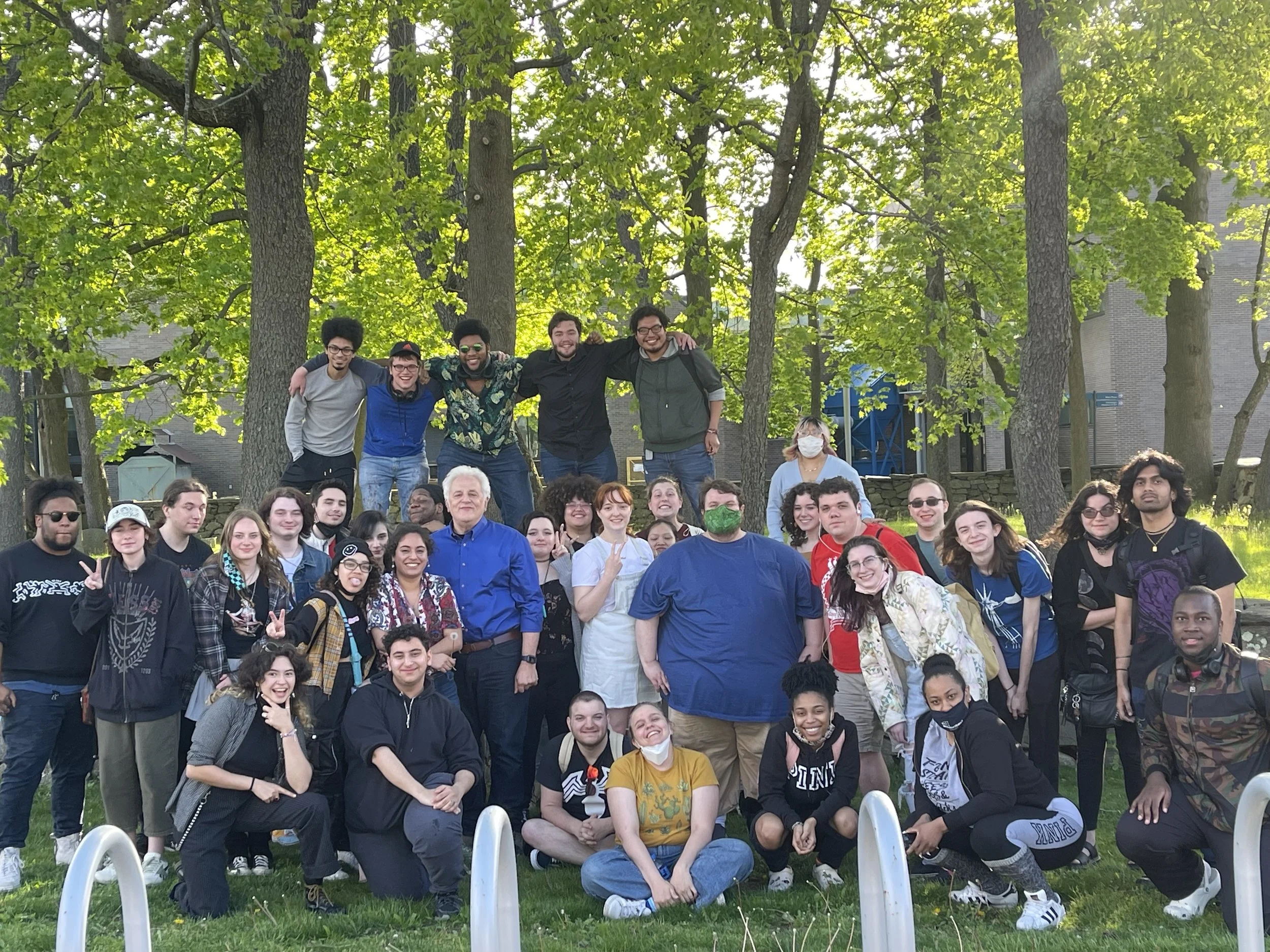A large group of diverse people gathered outdoors in a park with tall green trees, posing for a group photo. Some people are smiling, some are making peace signs, and a few are wearing masks. There are wide smiles and casual clothing among the group members.