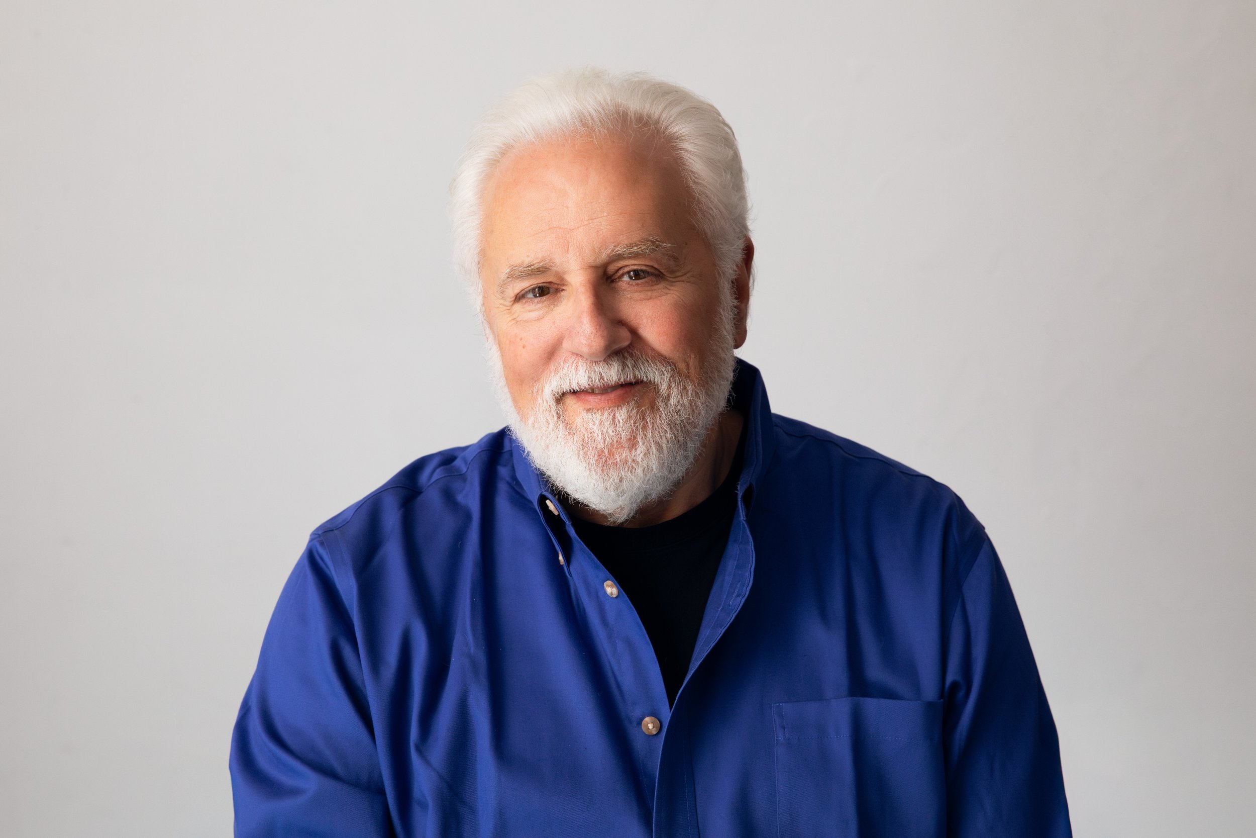 Close-up portrait of an elderly man with white hair and a beard, smiling slightly, wearing a blue button-up shirt against a plain light gray background.