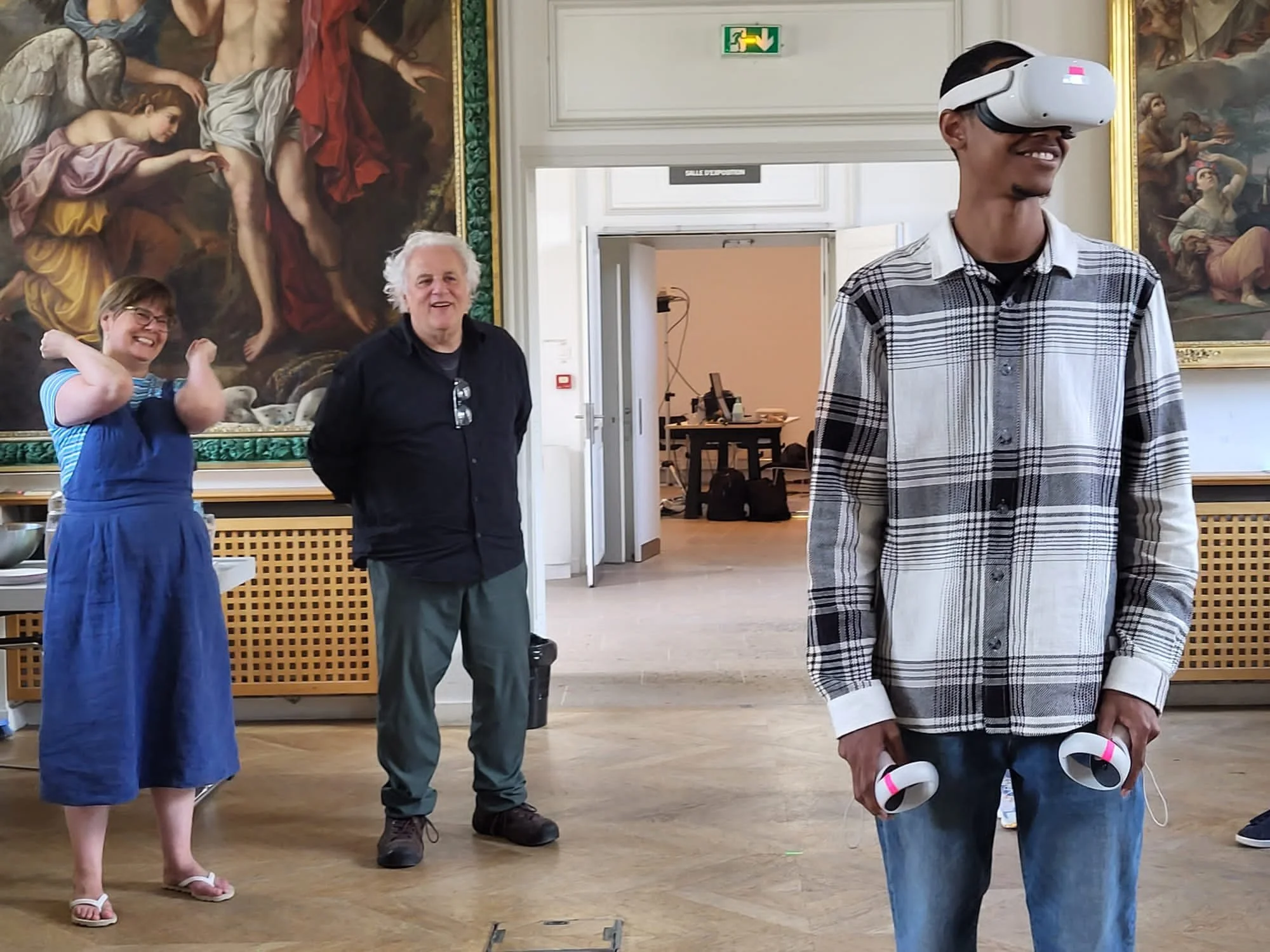 A group of three people standing inside a room with large paintings on the wall behind them. One person is wearing a virtual reality headset, smiling, and holding VR controllers. Two other individuals are standing nearby, smiling and watching, with one person wearing glasses and a striped shirt, and the other with white hair and a black shirt.