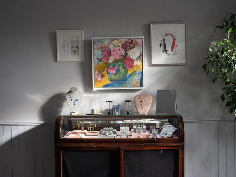 Jewelry display case with necklaces, rings, and earrings inside. Three framed artworks on the wall behind the case, including a floral painting in the center, a minimalistic face on the left, and an abstract figure on the right. A mirror and a potted plant are also visible.