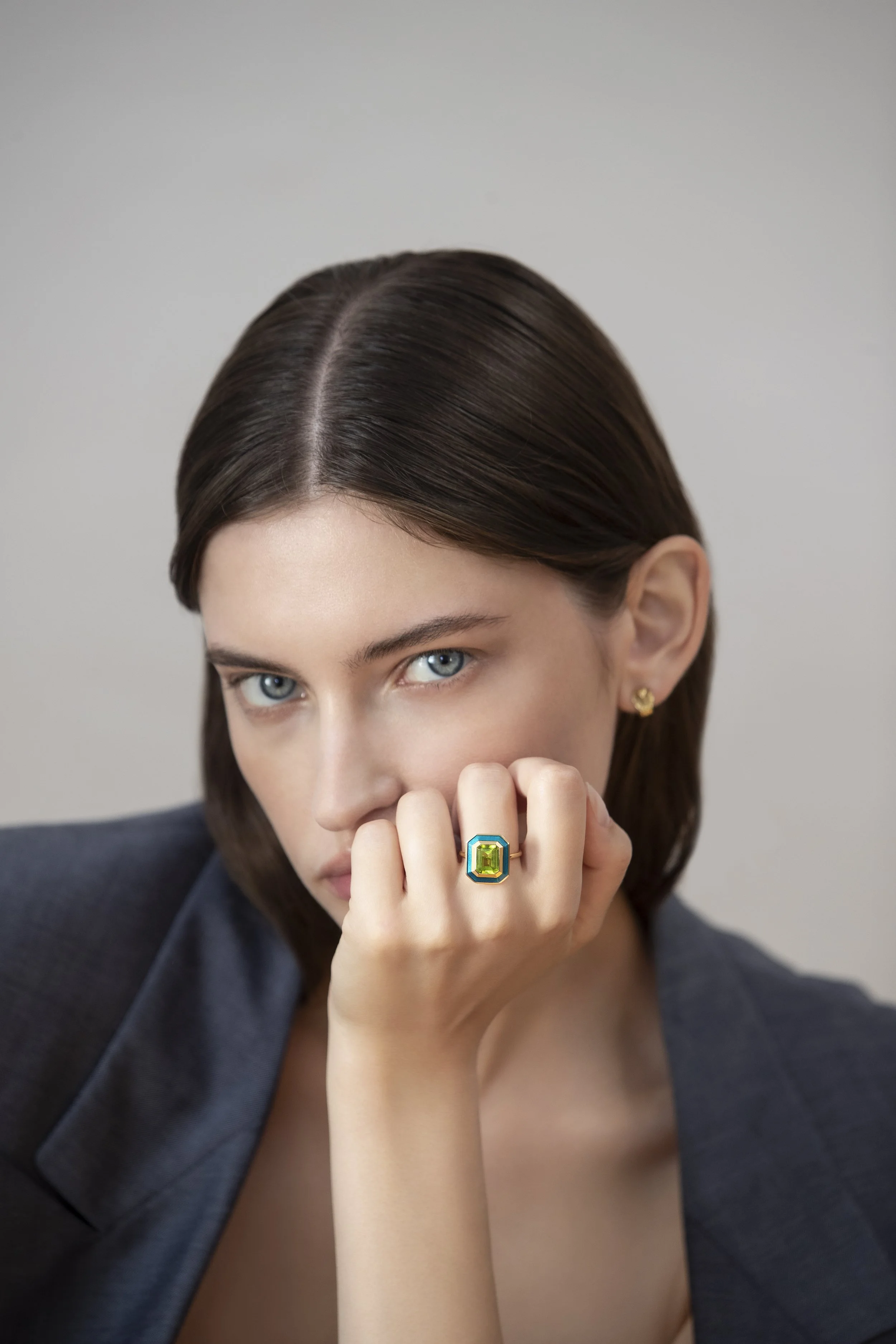 A woman with blue eyes and dark brown hair wearing a blazer, with her hand near her face showing a large green gemstone ring.