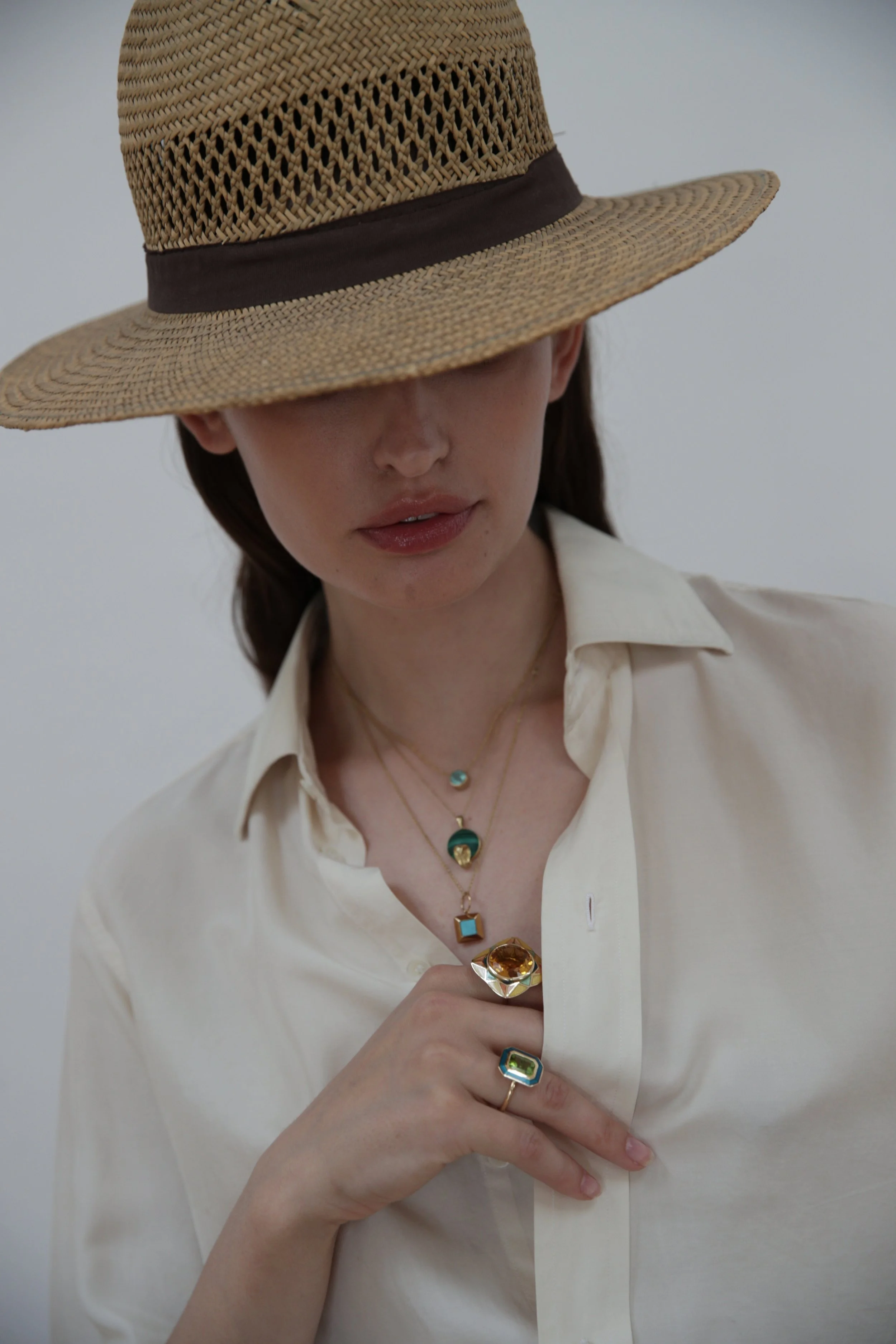 A woman wearing a wide-brimmed straw hat with a brown band, a cream-colored button-up shirt, and layered colorful jewelry, by Tarra Rosenbaum including necklaces and rings, against a plain light background.