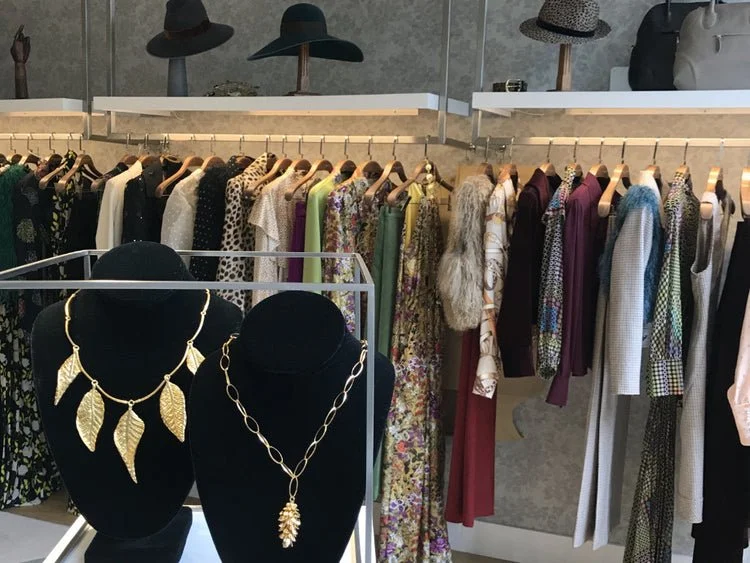 Display of gold jewelry necklaces on black bust stands in front of a clothing rack filled with colorful dresses and tops in a boutique store.