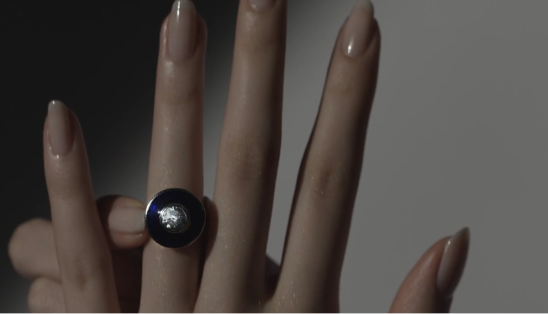 A hand wearing a black ring with a large clear gemstone, with the fingers slightly spread against a neutral background.