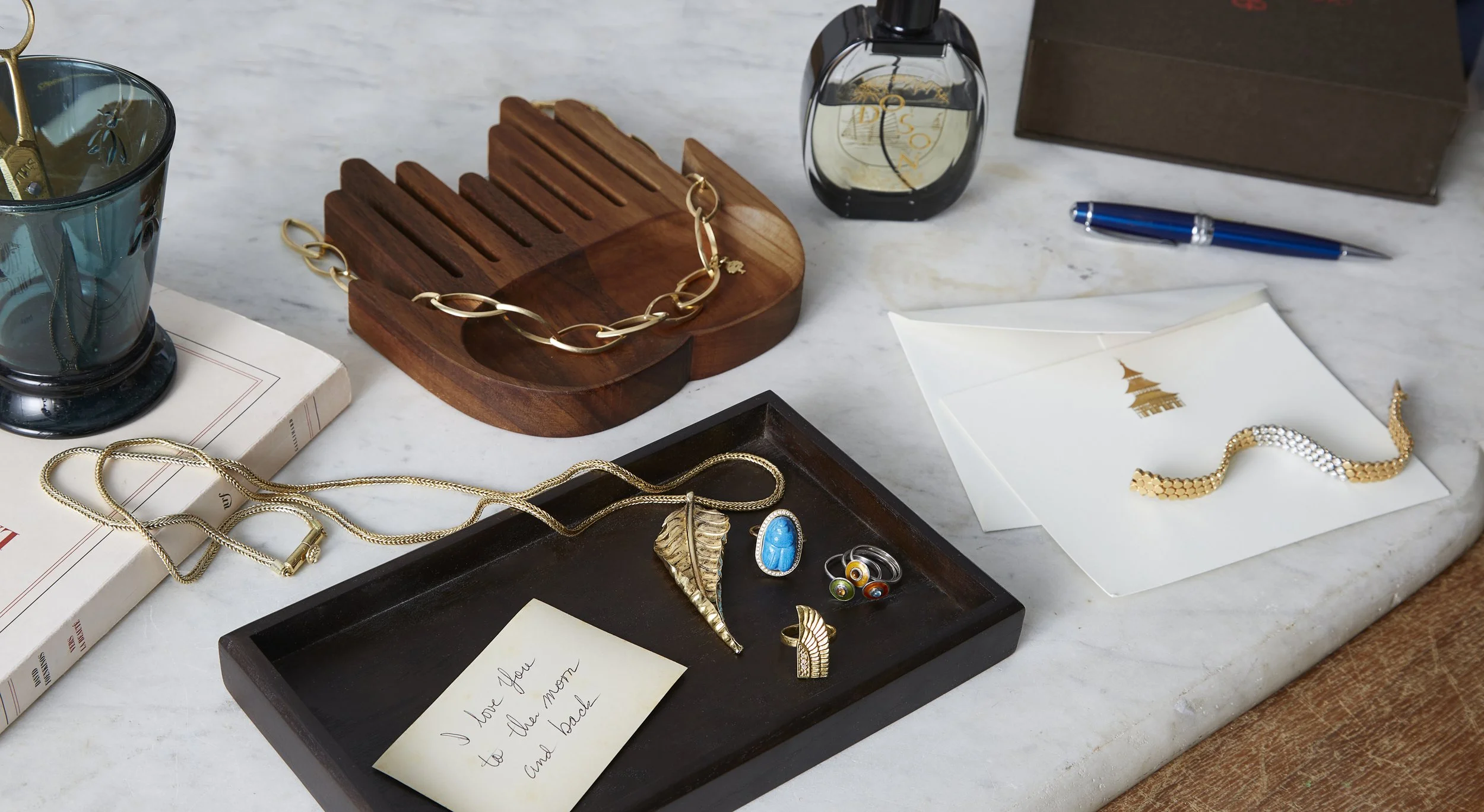 A collection of jewelry and decorative objects on a marble surface, including gold and silver necklaces, rings, a gold bracelet, a blue glass, a pen, and a letter with gold-embellished paper, with a jewelry box and a wooden jewelry display.