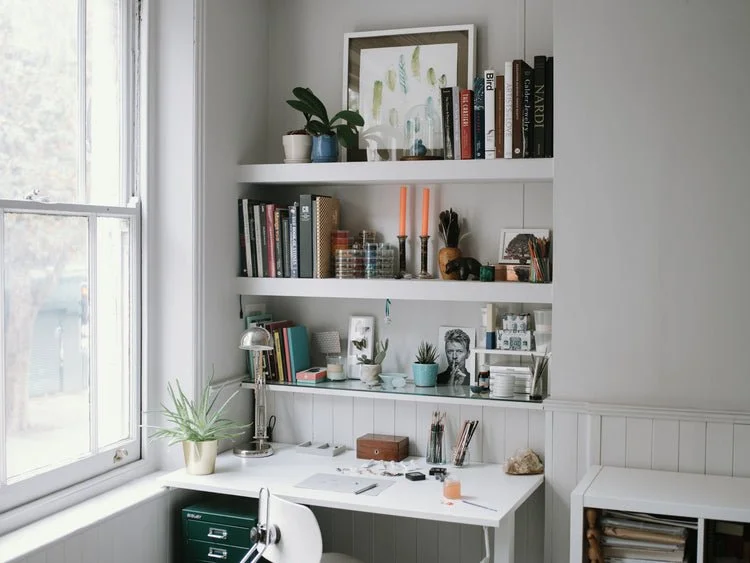 Home office nook with white desk, potted plants, bookshelves filled with books and decor, and a window letting in natural light.