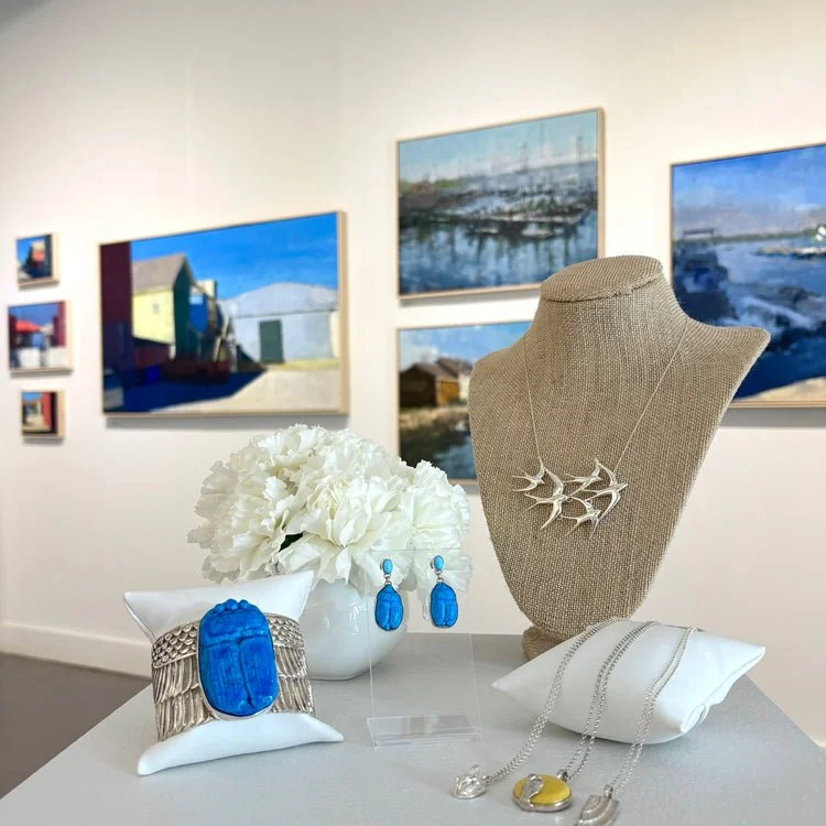 Jewelry display with a necklace featuring a silver branch design, matching earrings, a large blue ring, and a smaller blue ring, all arranged on a white table. A beige jewelry bust shows a silver necklace. A white vase with white flowers is also on the table, with artwork featuring beach and harbor scenes hanging on the gallery wall in the background.