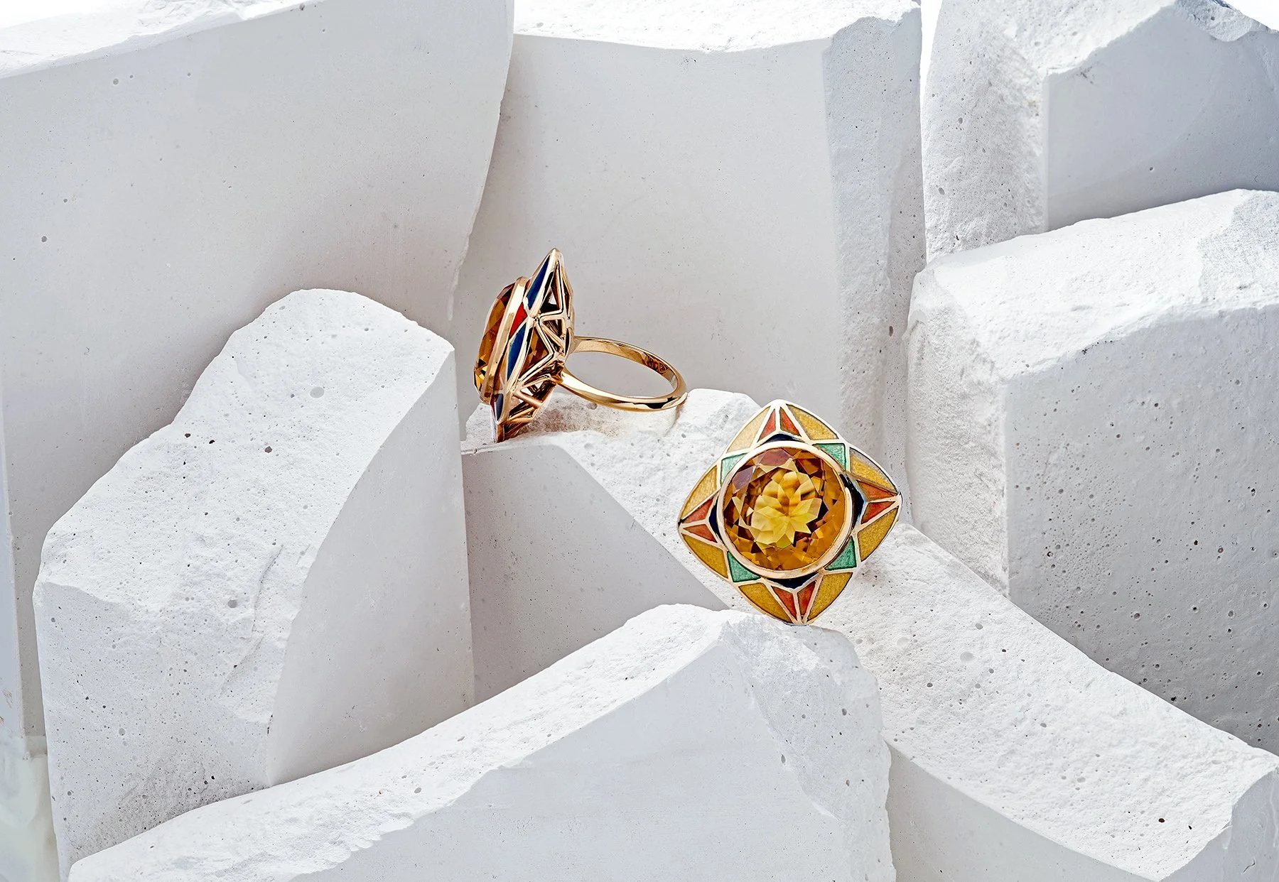 Two ornate jewelry rings with colorful geometric designs and a large yellow gemstone, displayed on white textured stone blocks. SUNRISE & SUNSET AURORA RING BY Tarra Rosenbaum. Gold ring with a large, faceted yellow citrine and vitreous enamel.
