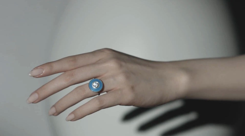 Close-up of a hand wearing a ring with a round blue bezel and a clear gemstone in the center, set against a plain background.