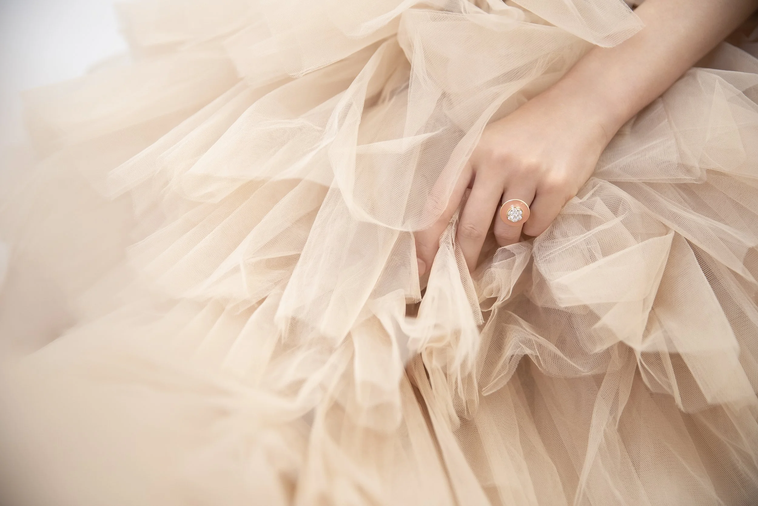 Close-up of a woman's hand wearing a large diamond ring, resting on a flowing, peach-colored tulle fabric.