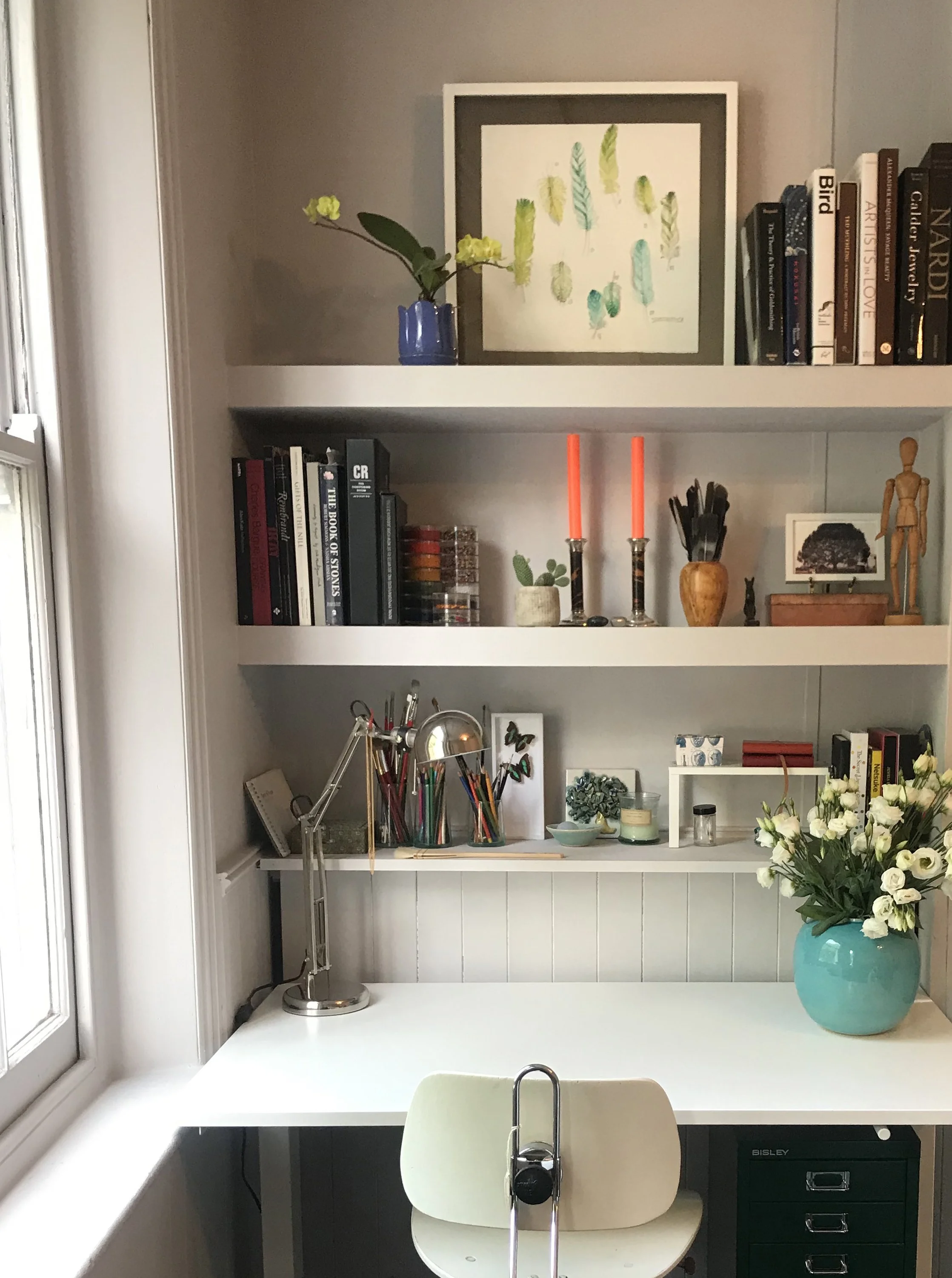 A white desk with a white chair facing a set of white shelves filled with books, art supplies, and decorative items, including a blue vase with yellow flowers on the top shelf, and a large white and blue flower vase on the right side of the desk.