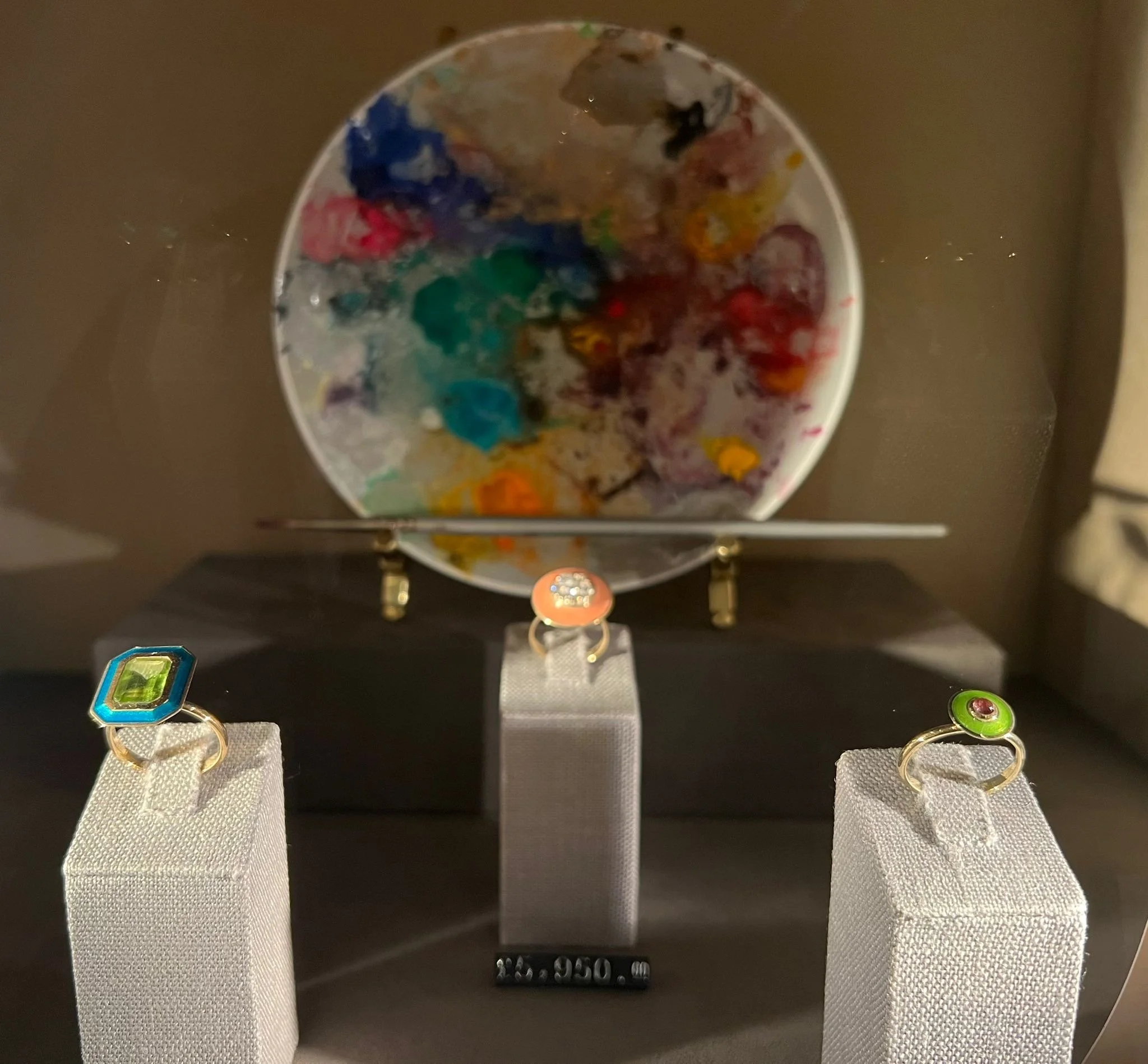 Display of three rings with colorful gemstones on white stands, set against a backdrop of a paint palette with mixed colors.