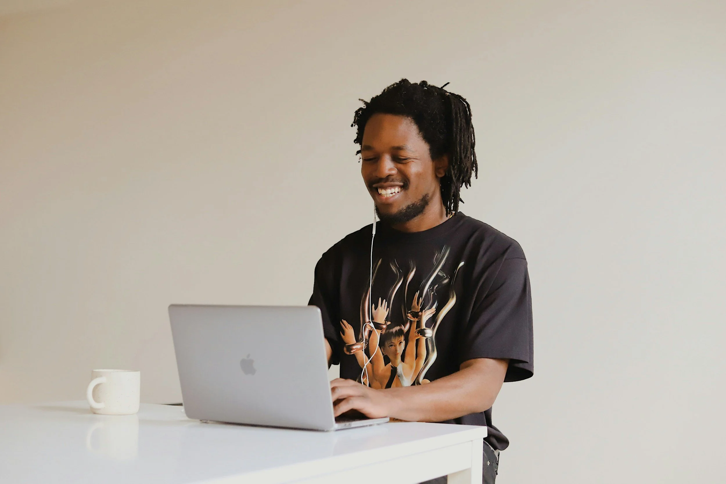 A man with dreadlocks smiling while working on a laptop at a white desk, with a white coffee mug to his left and a plain light-colored wall in the background.