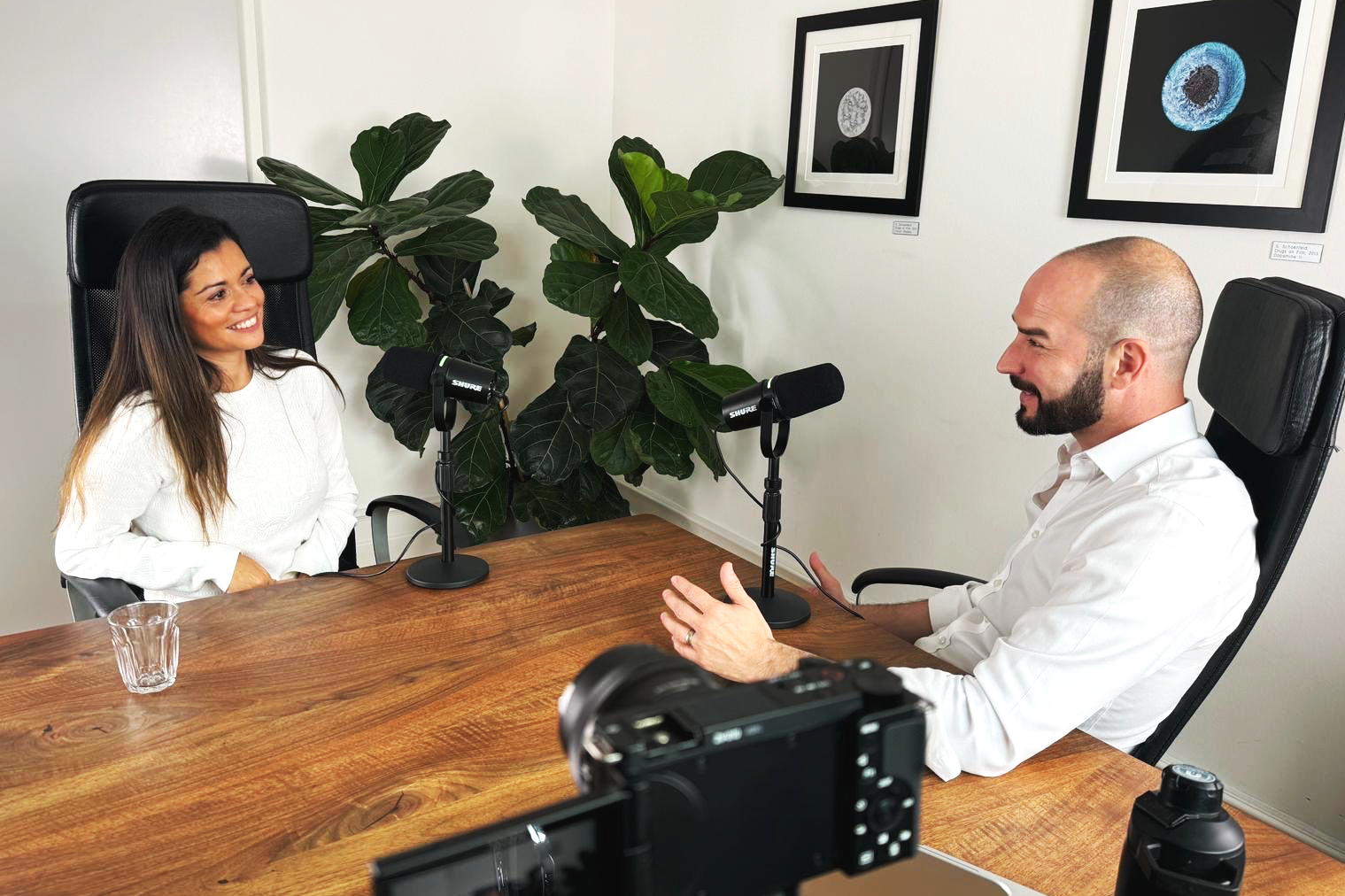 A woman and a man are sitting at a conference table with microphones in front of them, engaging in an interview or discussion. The woman is smiling and wearing a white sweater, while the man is gesturing with his hands and has a white shirt. There is a camera in the foreground and framed artwork on the wall behind them, along with potted plants.