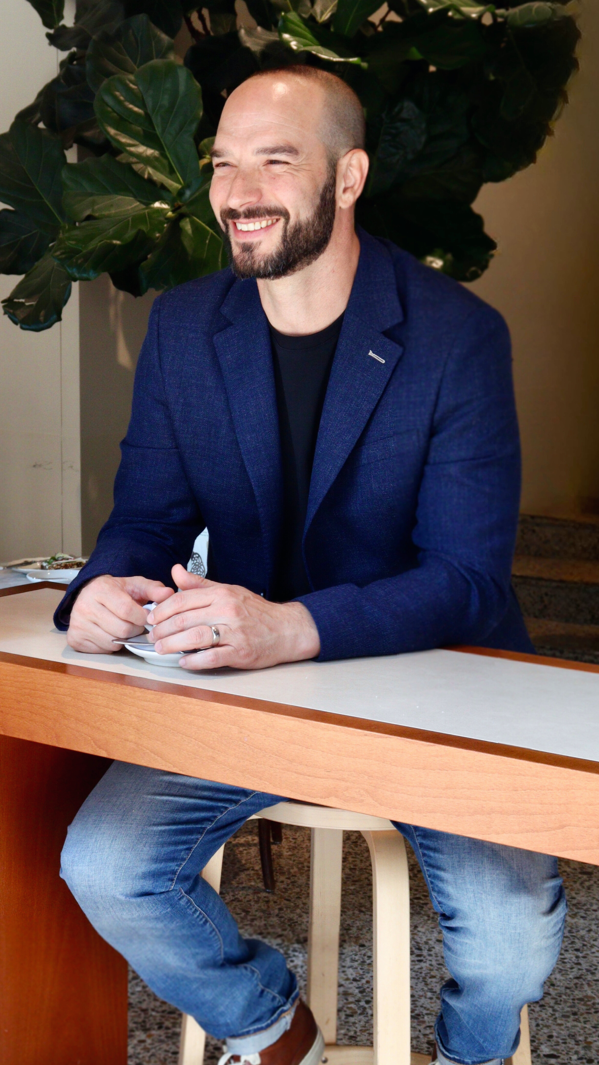 A man with a beard, smiling, sitting at a table with a plant in the background, dressed in a blue blazer, black shirt, and jeans.
