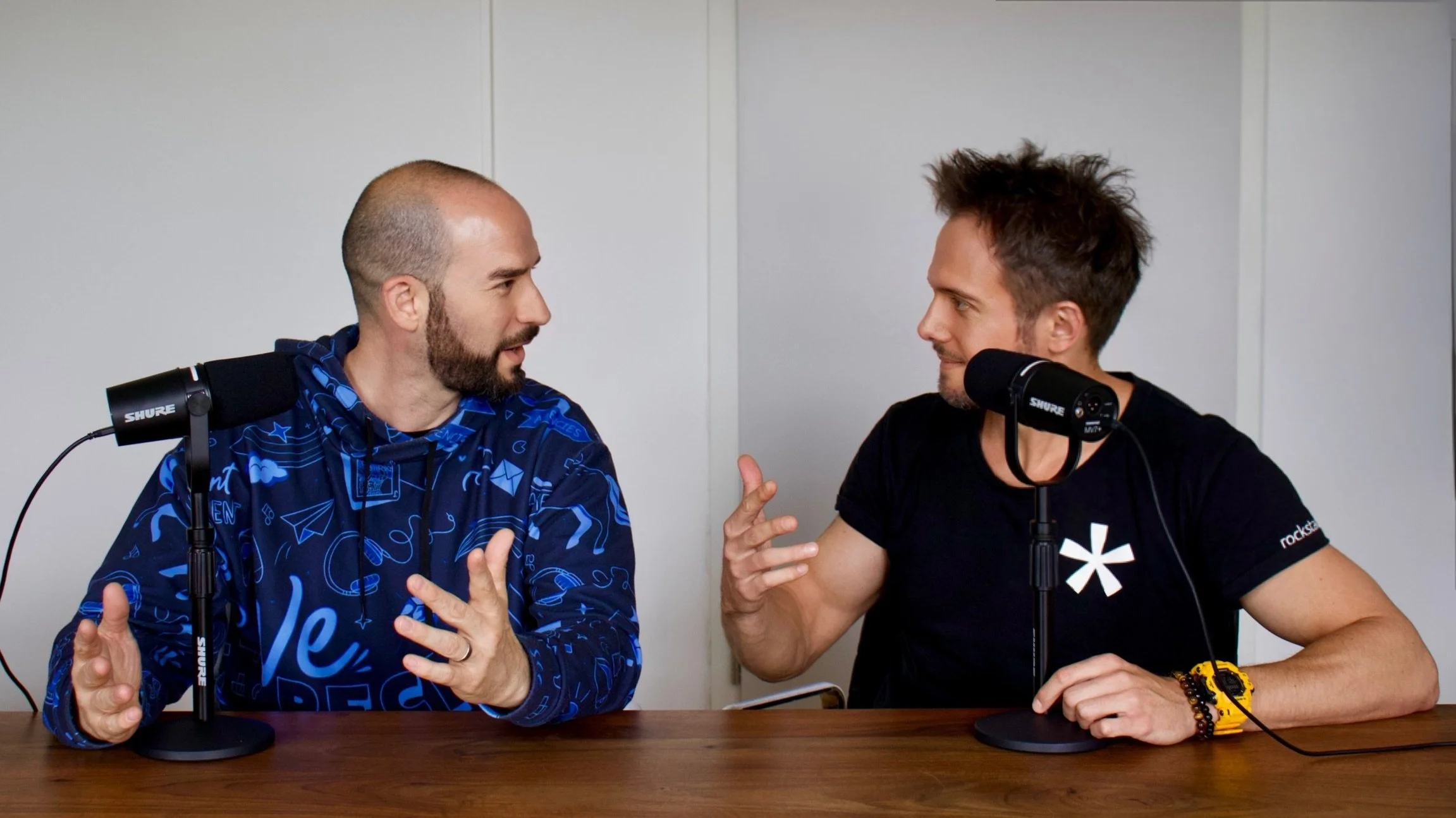 Two men are sitting at a table, speaking into microphones. They are engaged in a conversation and appear to be recording a podcast or interview. The man on the left is wearing a blue hoodie with graphic patterns, and the man on the right is wearing a black t-shirt with a white symbol. Both men are gesturing with their hands as they talk.