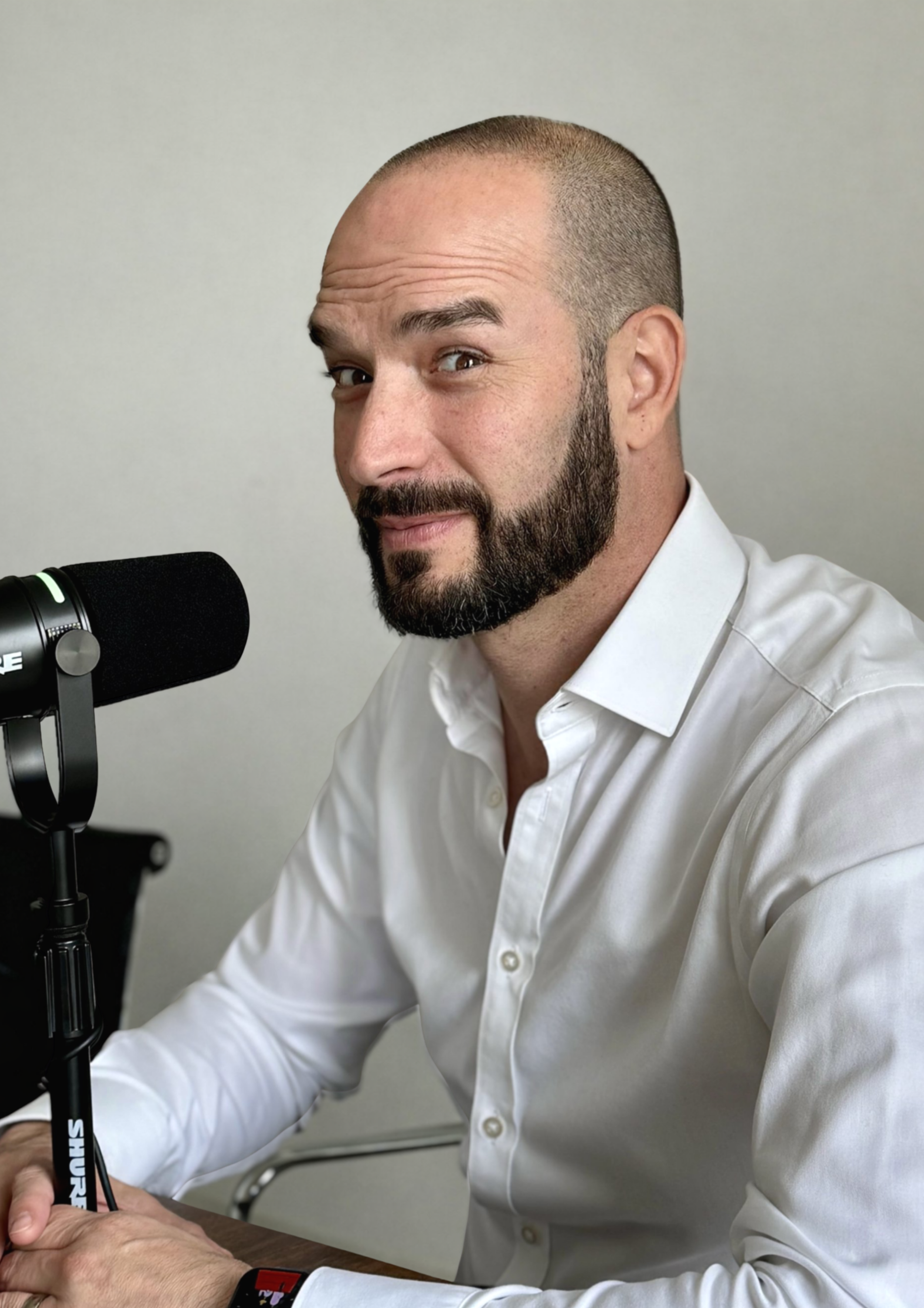 A man with a beard and a shaved head sitting at a desk in front of a microphone, wearing a white shirt.