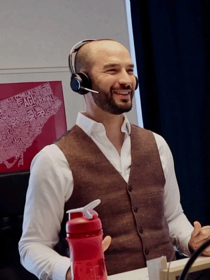 A man with a beard and shaved head wearing a white shirt and brown vest, smiling and wearing a headset, sitting at a desk with a red water bottle.