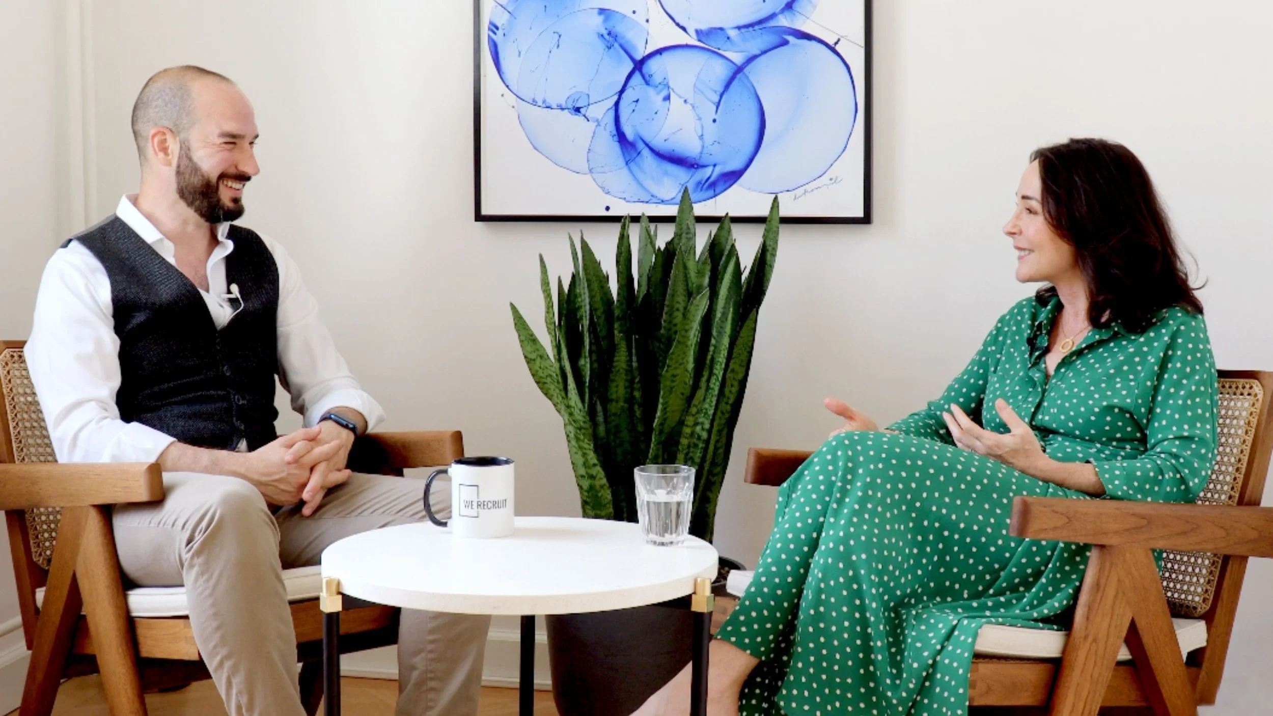 A man and woman are sitting in a conversation in a room with white walls and a piece of blue abstract art on the wall. A green leafy plant is between them on a small round table, which has a mug and a glass of water.