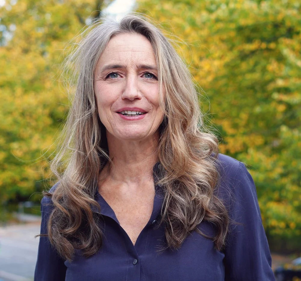 Eine ältere Frau mit langem, lockigem, grauem Haar steht outdoors vor einem Hintergrund mit grünen und gelben Blättern, trägt eine dunkelblaue Bluse.