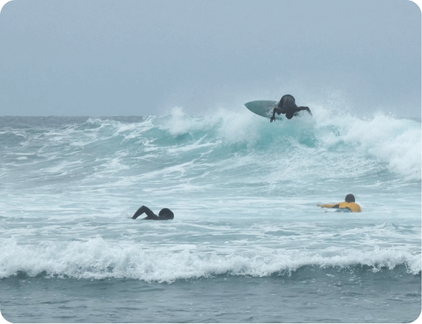 Time to Surf - Surfer mid-air performing a jump over a wave at the beach, with two other surfers floating on their surfboards nearby.